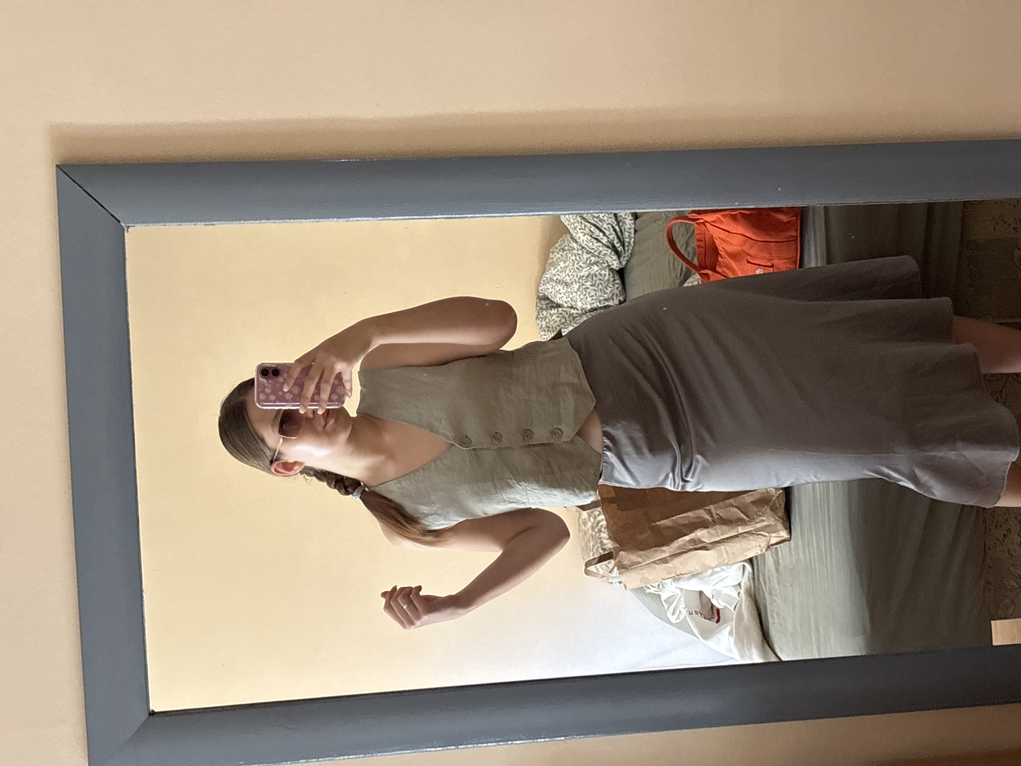 An indoor mirror selfie captures a young person, likely female, dressed in a casual outfit. The image itself is oriented vertically, showing a mirror with a dark grey frame against a light, warm yellow-beige wall.
The subject is seen from the chest down to the mid-thigh. She holds a purple-cased smartphone to take the picture, partially obscuring her face. She wears a light olive-green sleeveless vest-style top with four small buttons down the front, paired with a long, flowing dark grey skirt. Her long, dark brown hair is styled in a braid that rests over her left shoulder. She has sunglasses on her face, and a ring is visible on a finger of her left hand. Her expression is not discernible.
Reflected in the mirror behind the subject, glimpses of the room are visible. These include a bed or sofa covered with a greenish-white patterned duvet or blanket, an orange bag with handles, a crumpled brown paper bag, and a white piece of fabric or garment with partial text "an" and "dy" visible. Another section shows a plain dark green sheet or fabric.
The scene depicts a personal moment, likely in a bedroom or dressing area, where the subject is showcasing her attire. The overall setting is a private interior space in Bečići, Montenegro.