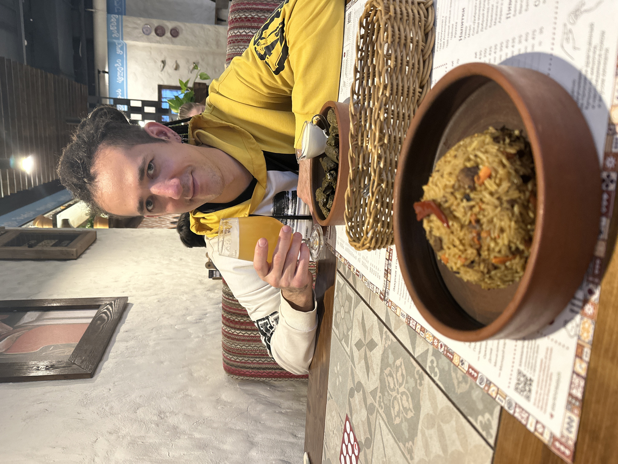 The image captures a man seated at a table in what appears to be a Georgian restaurant. The man, with dark, curly hair and ear piercings, wears a yellow hooded sweatshirt over a white long-sleeved shirt and looks towards the camera with a slight smile. He holds a glass of light-colored beverage, possibly beer or juice.
On the wooden table, which features a central section of patterned tiles, two traditional dishes are present. One is a rustic brown bowl filled with pilaf, a rice dish with visible meat and vegetables. The other is a bowl of dolma (stuffed grape leaves), over which a small pitcher is pouring a white sauce, likely yogurt or sour cream. A wicker basket and a menu are also on the table.
The background shows a light-colored, textured wall with framed artwork and some indoor plants. Behind the man, a patterned cushion or upholstery suggests booth seating. The lighting is soft, indicating an indoor evening setting.
Notable text includes Georgian script on the wall, reading "საქართველო იყო და იქნება," which translates to "Georgia was and will be." The menu on the table displays Russian text, prominently featuring "Напитки" (Drinks) and a QR code, confirming the restaurant's location in Russia, specifically Saratov as contextually provided. The man appears relaxed and content, enjoying his meal.