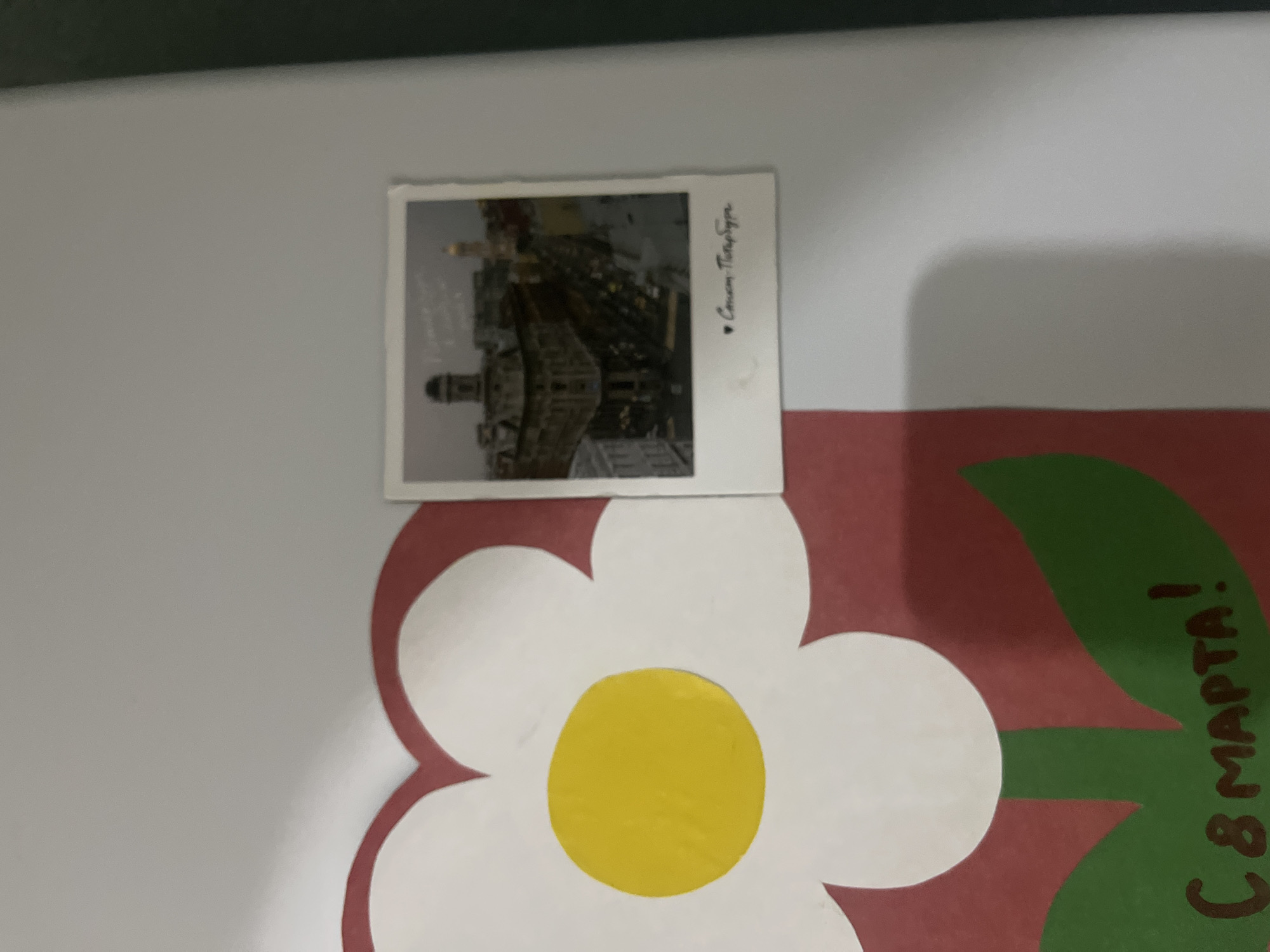 The image displays a portion of a wall decorated with a large white and red flower illustration. Attached to the wall, to the right of the flower, is a small polaroid-style photograph featuring a cityscape. The photograph shows buildings with architectural details and a dome, suggesting an urban European setting. Below the cityscape in the photograph, handwritten text in Cyrillic script is visible, which appears to read "С любовью - Петербург" (With love - Petersburg). The bottom portion of the flower illustration shows the text "С 8 МАРТА!" (Happy 8th of March!) also in Cyrillic. The overall context suggests a celebratory decoration, possibly for International Women's Day, with a personal touch referencing St. Petersburg. The image does not contain any information to suggest the location is Gorodets, Russia.