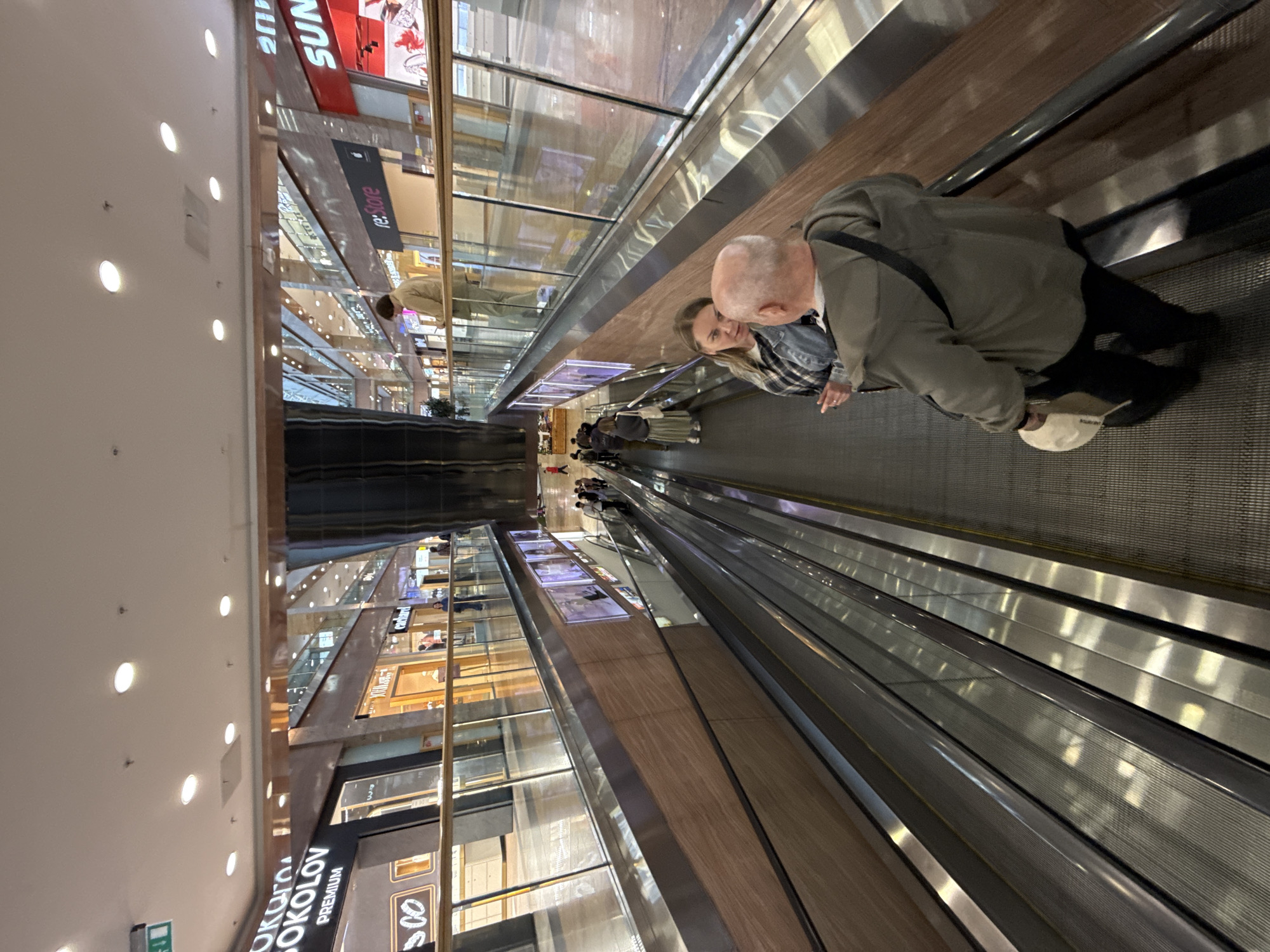 The image captures a view from an escalator in a multi-level indoor shopping mall. On the escalator in the foreground, a man with a shaved head, wearing a grey hooded jacket and black pants, is walking down while carrying a light-colored hat in his left hand. To his right and slightly behind him, a woman with blonde hair, wearing a denim jacket over a plaid shirt and dark pants, is looking towards the man. They appear to be engaged in conversation. Other shoppers are visible on the escalator further down, as well as on the walkways of the different levels of the mall. The mall features storefronts with illuminated signs, including "SOKOLOV" and "re:Store". The architecture includes wooden paneling and glass railings. The overall environment suggests a modern, well-lit commercial space.