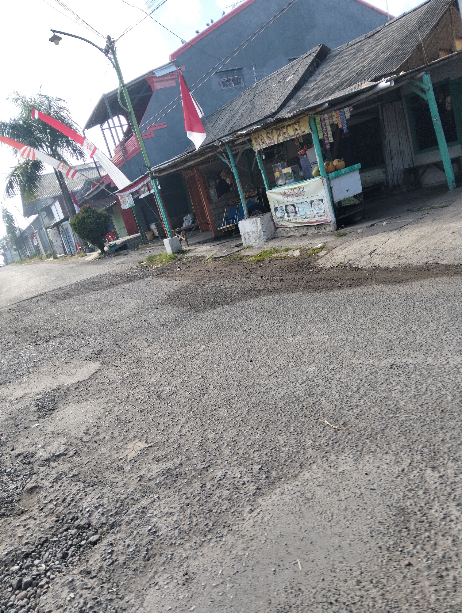 The image depicts a street scene in Muncar, Indonesia, characterized by a weathered, gray asphalt road with patches of damage and scattered pebbles. To the right of the road, a small roadside stall with a corrugated metal roof is visible. This stall appears to be a food vendor, indicated by signage that reads "NASI PECEL" (Peanut Sauce Rice) and "WARUNG" (small shop). The stall displays various packaged goods and food items, with some coconuts or similar round produce on a counter. In front of the stall, a banner features images of food and text, including "NASI BEBEK," "MIE SEDAP," "ES DEGANA," and "ES CAMPUR," suggesting a variety of menu options. A wooden bench is placed outside the stall.
Behind the stall, a two-story building with a dark gray facade and red accents can be seen. The building has a pitched roof and visible windows. Further down the street, other buildings and structures are partially visible, suggesting a residential or commercial area. Red and white Indonesian flags are displayed on poles and draped across the street, hinting at a national celebration or holiday. A street lamp is mounted on a pole on the left side of the road. A small, trimmed green tree stands near the stall. The overall impression is one of a typical, somewhat aged, street-level commercial area in a town or city in Indonesia.