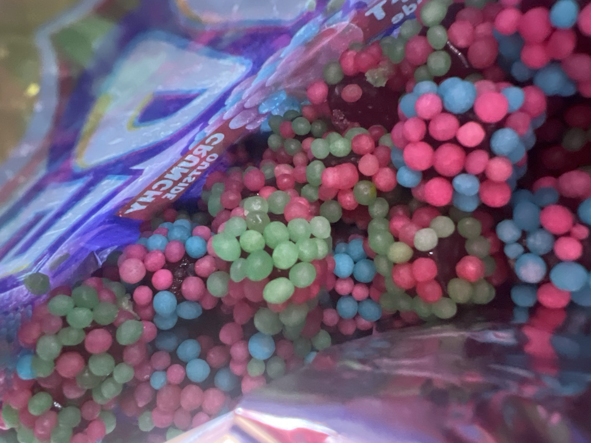 The image shows a close-up view inside a bag of colorful candy. The candy pieces are small, spherical, and come in bright shades of pink, blue, and green. Some of the candy pieces are clustered together, while others are scattered. The bag itself appears to be made of a reflective, possibly plastic material, with some purple and white branding visible. Text on the branding reads "CRUNCHY" and "OUTSIDE," suggesting the candy has a crunchy exterior. The lighting is bright and appears to be coming from above. The background is blurred and indistinct, but the focus is clearly on the candy. There is no information within the image to indicate the location in Eretria Municipal Unit, Greece.