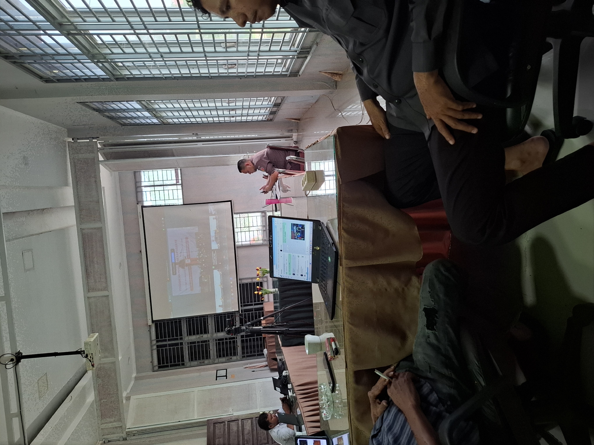 The image captures a meeting or presentation taking place in a room in Lhokseumawe, Indonesia. The room has a lattice-style ceiling, large windows with bars, and white walls.
Several individuals are present. One man sits near a projector screen, which displays a presentation. A laptop is on the table in front of the screen. Other people are sitting around tables, some of whom are partially visible in the foreground of the image. One person appears to be smoking.
A video camera on a tripod stands between the presenter and the screen. There are also decorative vases with flowers on the table. Glasses of water are set up.