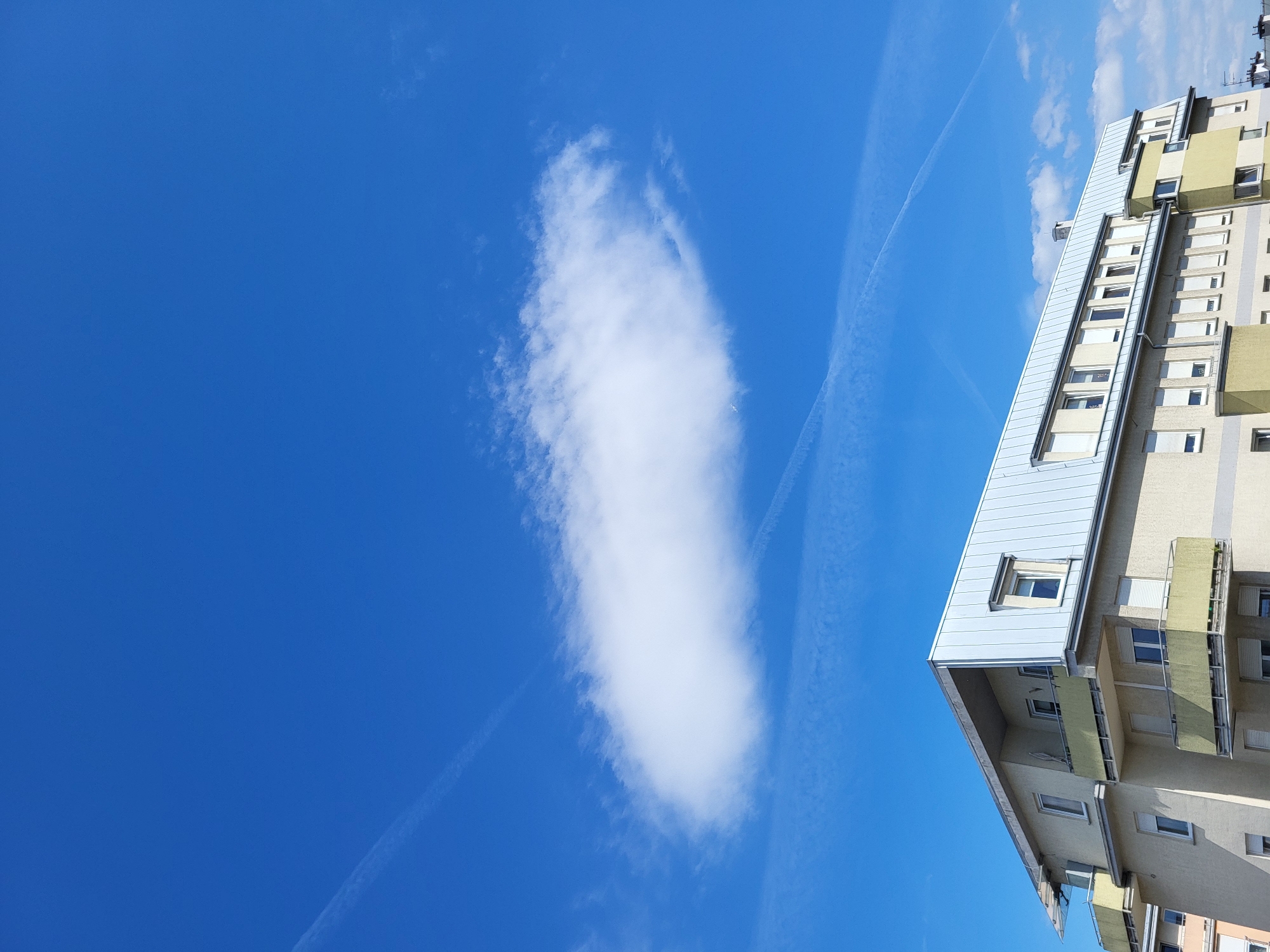 The image depicts a view of a building against a bright blue sky with a single elongated white cloud and contrails. The building is light-colored with multiple windows and balconies, and it appears to be an apartment or office complex. The sky is clear except for the prominent cloud and the contrails left by aircraft. The photo is taken from a low angle, making the building seem taller. The location details are unavailable.