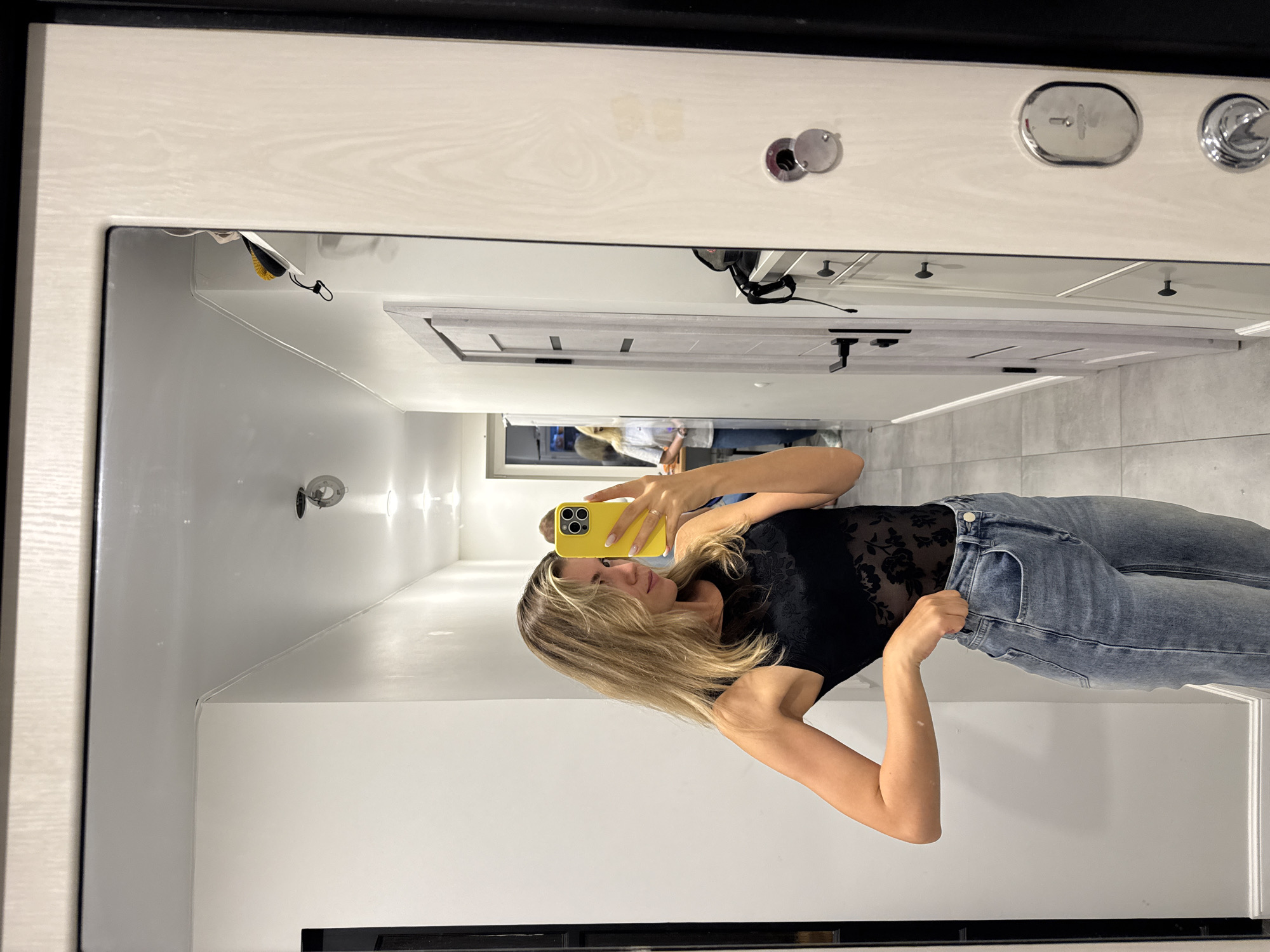 A young woman with blonde hair takes a mirror selfie in a hallway. She is wearing a black lace bodysuit and light-wash blue jeans. The hallway appears to be in a residential building, with light gray tiled floors and white walls. To the right of the woman is a large closet door with a wood-grain finish. In the reflection, another woman can be seen standing further down the hallway. The overall lighting is bright, suggesting the photo was taken during the day or in a well-lit interior space.