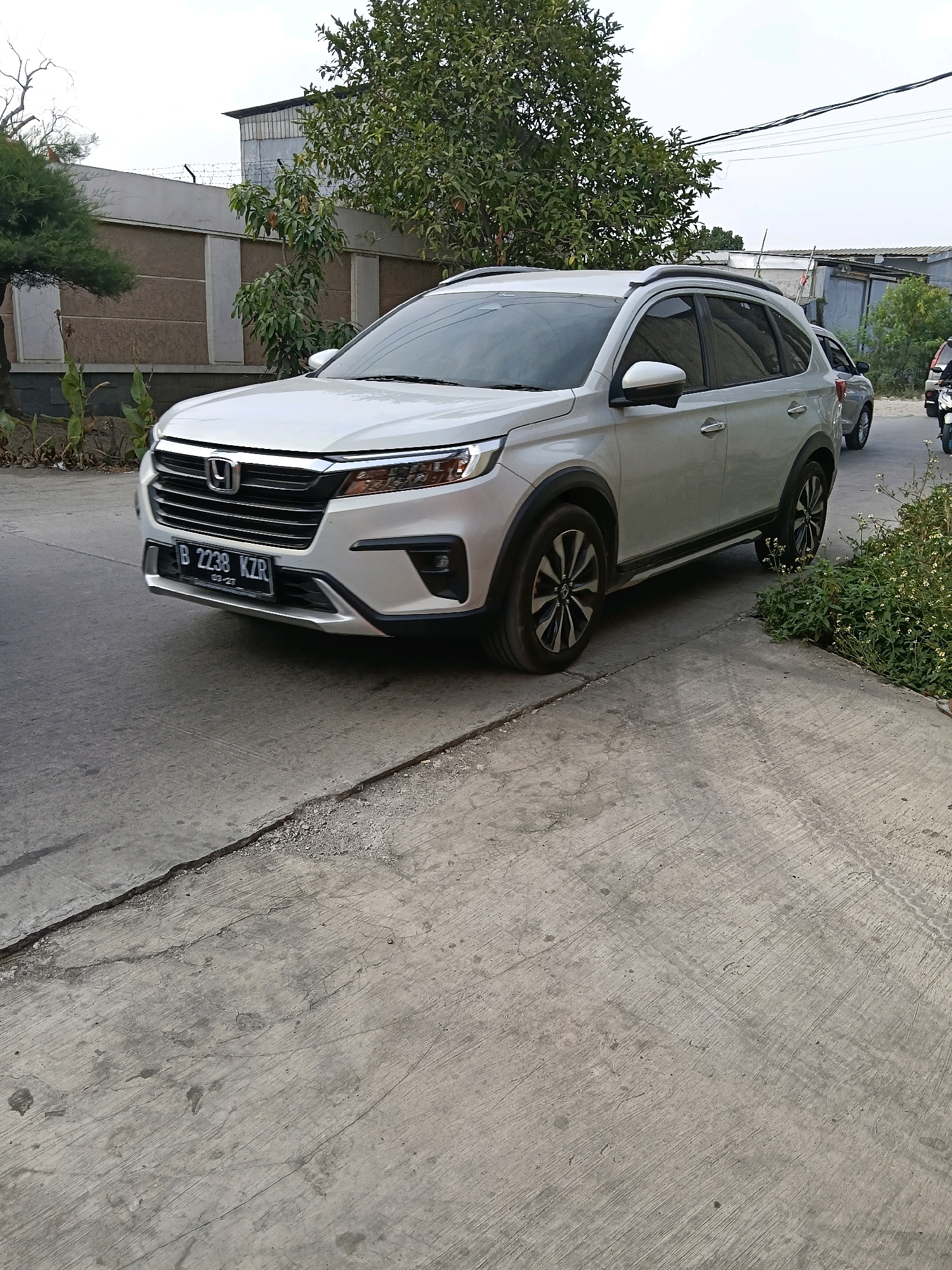 A silver Honda BR-V SUV with license plate B 2238 KZR is parked on a concrete road in the Belimbing area of Indonesia. The vehicle is facing towards the viewer's left, with its front end slightly angled away. The SUV has a silver grille with the Honda logo, and black accents around the fog lights. The wheels are alloy with a multi-spoke design. A growth of small, white-flowered plants is visible to the right of the vehicle. In the background, a concrete wall with a barbed wire fence runs along the property line. Behind the wall, there are trees and buildings. The sky is overcast.