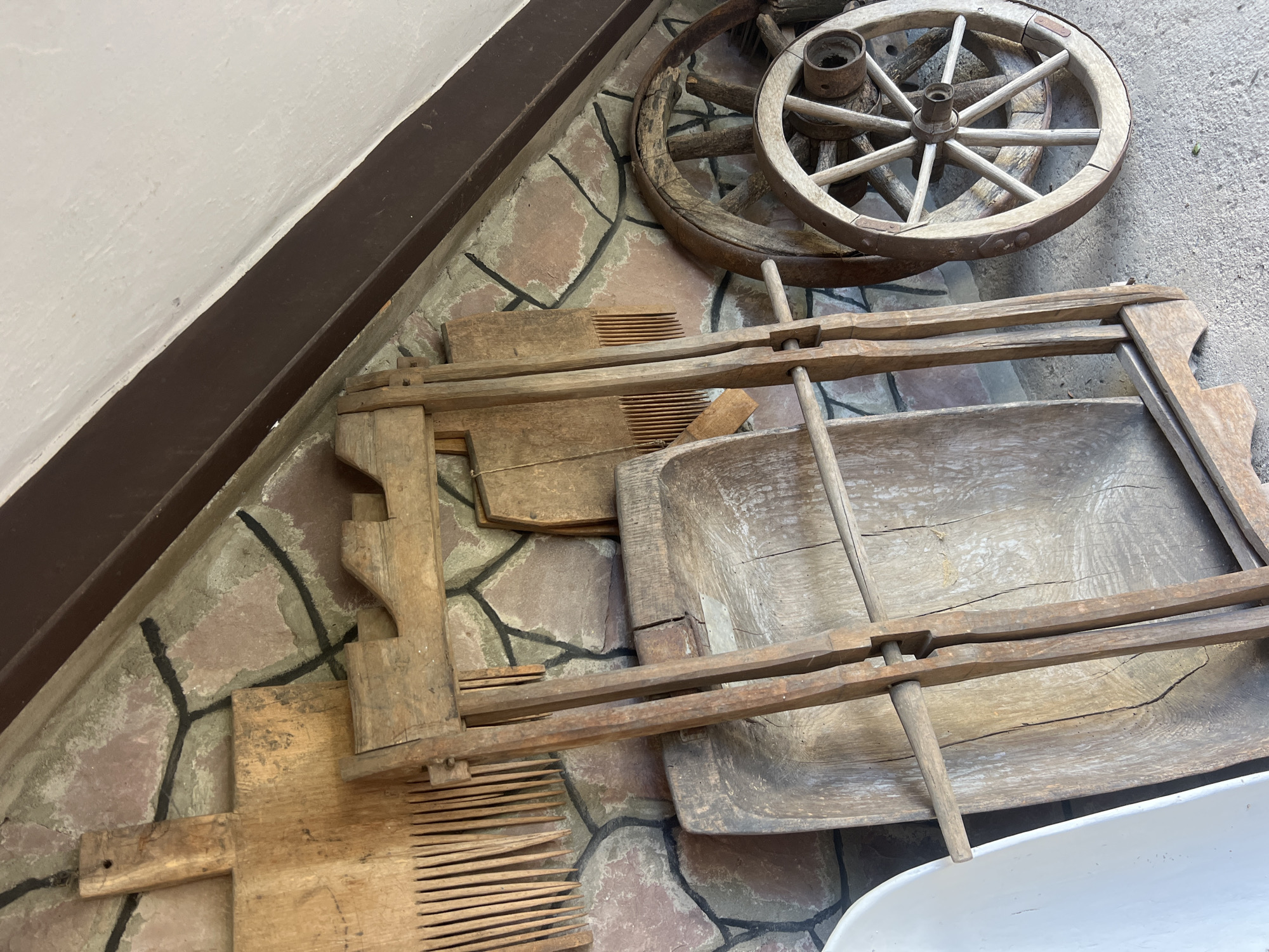 In Yaremche, Ukraine, a collection of antique agricultural tools is displayed against a stone wall. Prominently featured are two wooden wagon wheels, one larger and darker than the other. To the left of the wheels, a wooden, foldable contraption with two upright supports and a horizontal rod is positioned. Behind this, a large, weathered wooden trough or basin is visible. To the far left, a portion of a white object, possibly a wheelbarrow, is present. Hanging on the stone wall are two wooden combs with numerous tines, and another wooden board. The scene is set outdoors on a concrete surface, under an overhang with a dark brown trim and a white ceiling. The stone wall is composed of irregularly shaped, pinkish-brown stones with dark grout lines. The overall impression is one of rustic history and traditional craftsmanship.