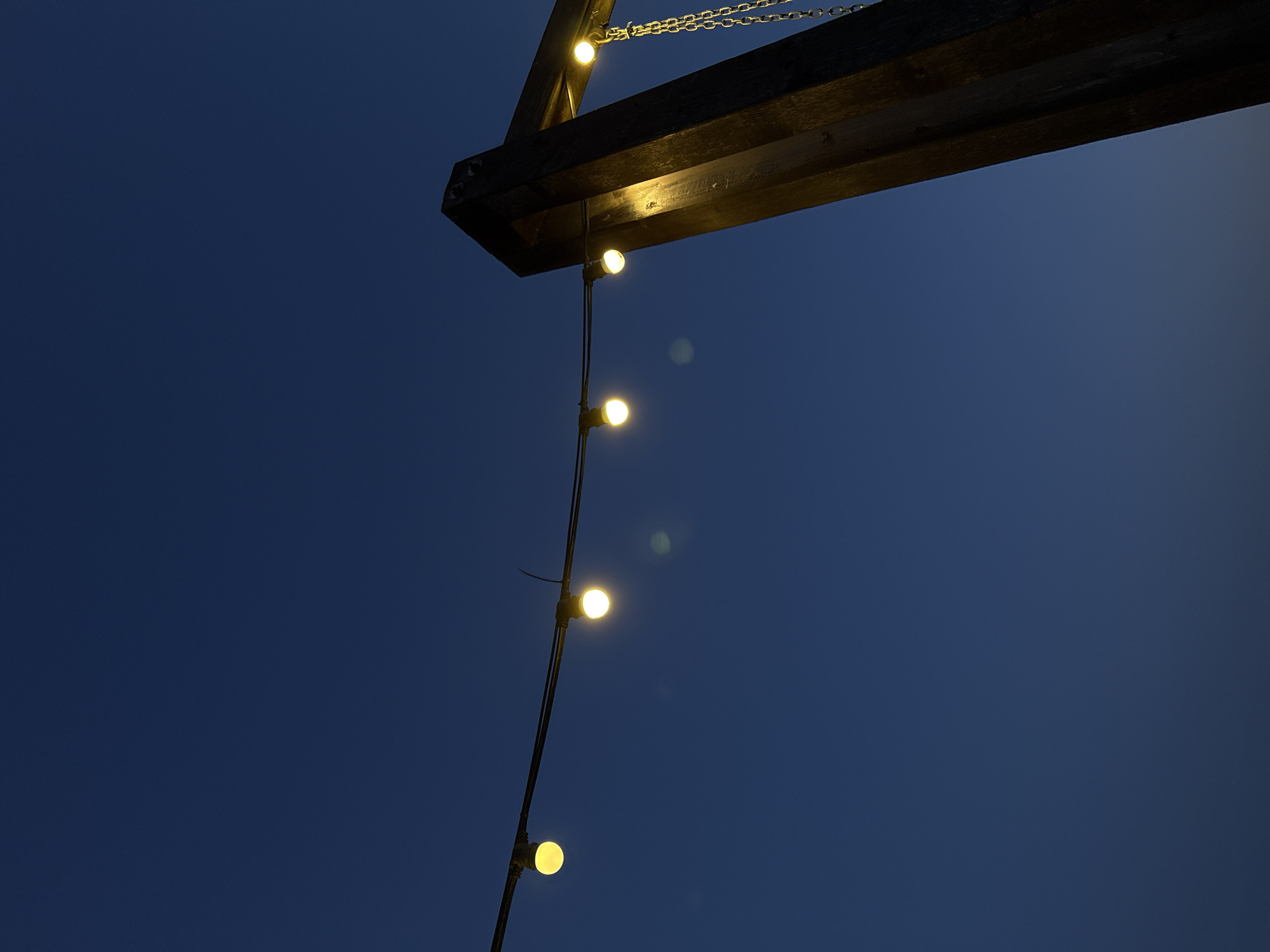 A low-angle shot captures a string of evenly spaced, illuminated round lights hanging from a dark wooden beam structure against a deep blue twilight sky. The beam is partially supported by a chain, suggesting it is an overhead fixture. The location is not identifiable from the provided image.