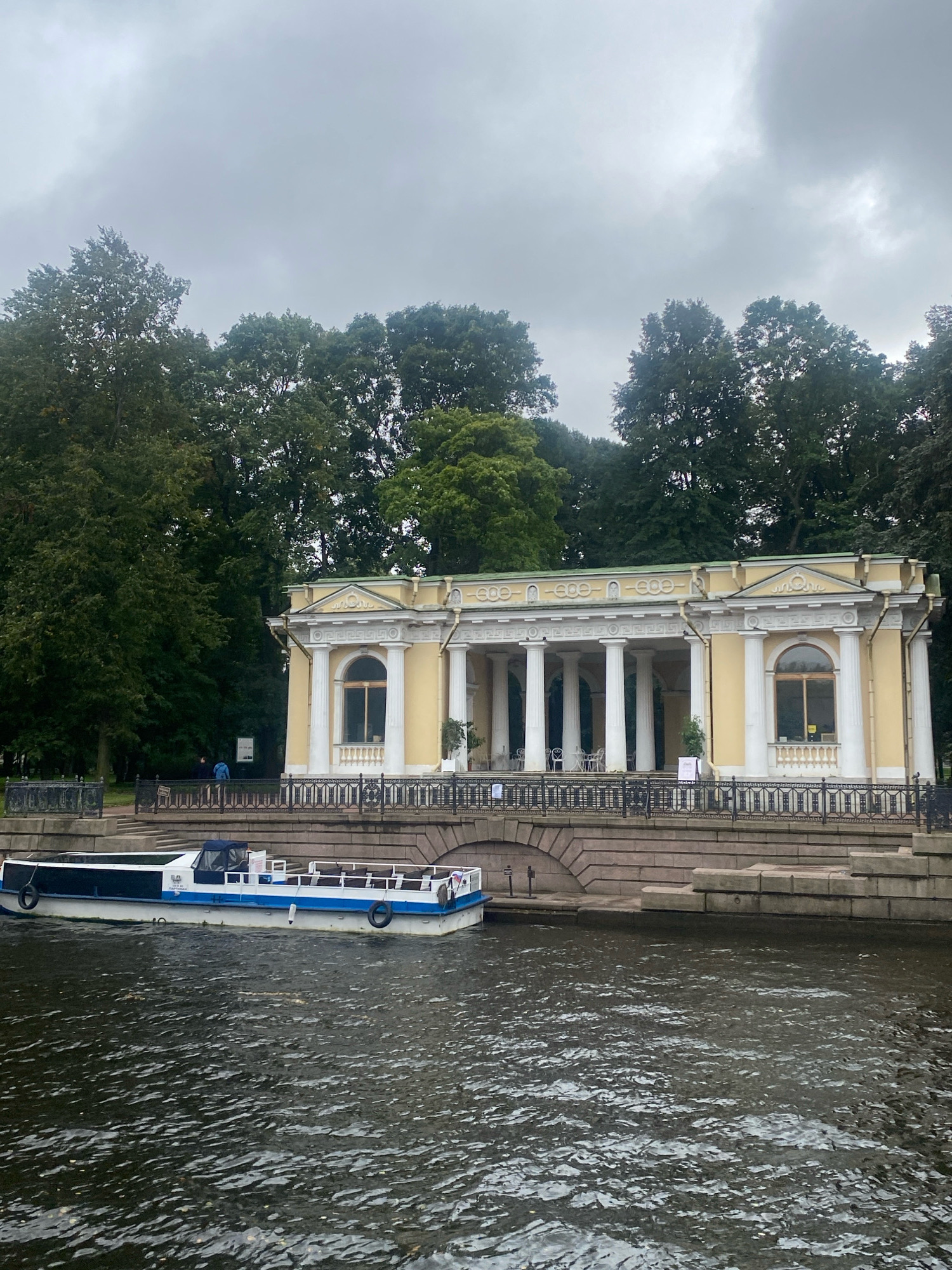 This is a full-shot, eye-level, outdoor photograph of a neoclassical pavilion on a cloudy day. The pavilion, painted a pale yellow with white columns and trim, features a portico with tall white columns, arched windows, and ornate decorative elements along the roofline. It is situated on an elevated embankment overlooking a dark, rippling body of water, likely a canal. A long, white and blue boat with a canopy is docked in the foreground on the water. Behind the pavilion, a dense line of dark green trees forms a backdrop. A black metal fence separates the pavilion from the embankment, and a few scattered figures are visible in the distance near the pavilion. The sky is overcast and gray, suggesting a cool or rainy atmosphere. The architectural style and the presence of a canal suggest this scene is in Saint Petersburg, Russia, potentially at a park or historical site like the Tauride Garden or Summer Garden.