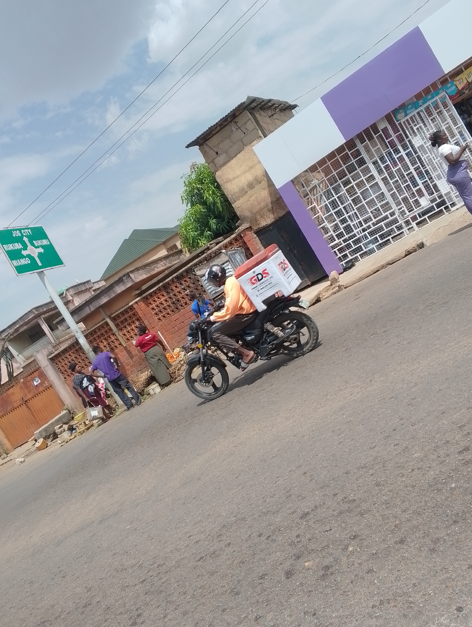 The image captures a street scene in Jos, Nigeria. A motorcycle, bearing a "GDS Prime Logistics Ltd" delivery box, is being driven on the road. The motorcycle rider is wearing an orange shirt and a helmet. In the background, there's a street sign indicating directions to "Jos City," "Rukuba," "Bukuru," and "Miango." Several individuals are present on the sidewalk near a building. One woman stands near a store with a white and purple facade. The sky is partly cloudy.