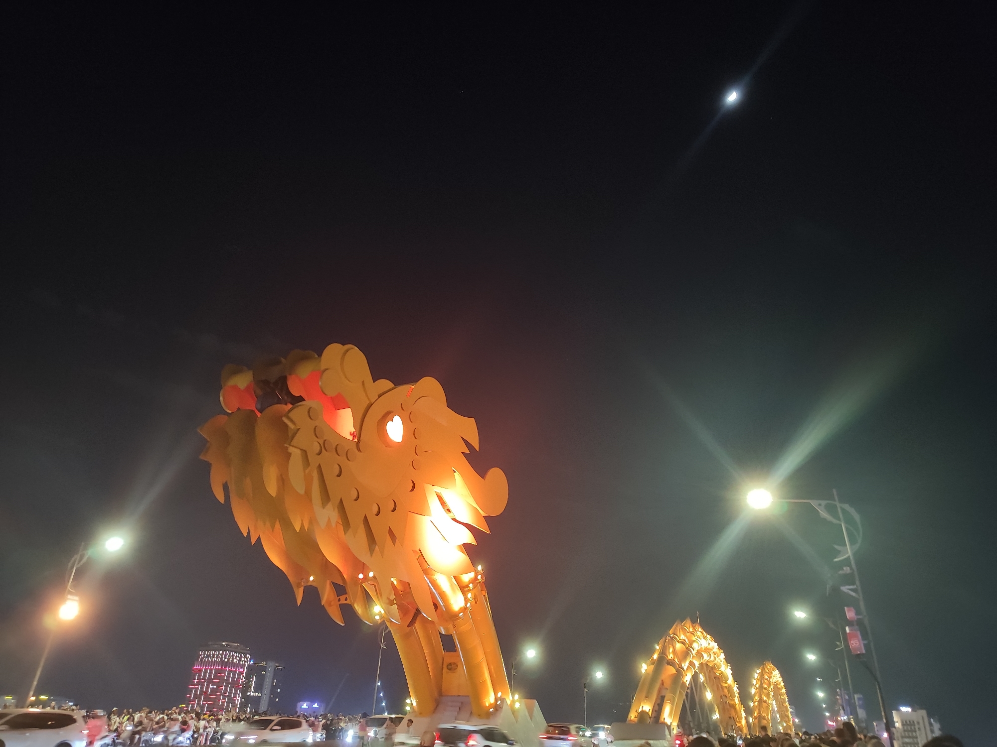The image depicts a nighttime scene in Đà Nẵng, Vietnam, featuring the illuminated Dragon Bridge. The bridge's dragon head is prominent, with lights giving it a golden appearance. The dragon head has red and golden lighting details. The scene includes other parts of the bridge, illuminated as well.
A crowd of people is visible near the bridge, along with cars and motorcycles suggesting traffic on the bridge and surrounding streets. Buildings are visible in the background, one of which appears to have a red, lit-up design. Streetlights are present, contributing to the overall illumination of the scene. The sky is dark, with a visible moon and possibly other faint stars.