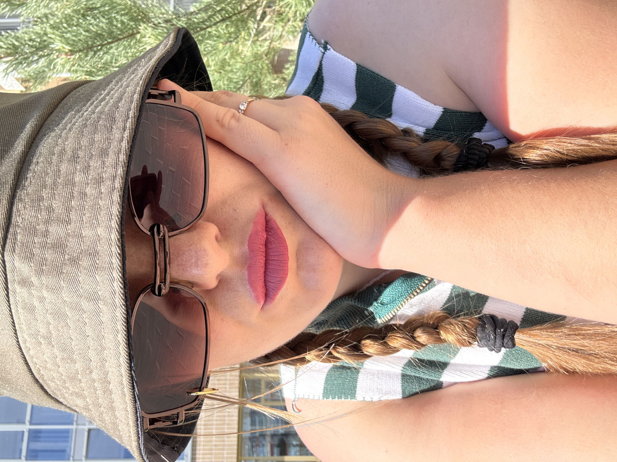 The image shows a close-up of a woman wearing a bucket hat and large sunglasses. She has her hand resting on her cheek, and her lips are painted with a pink lipstick. Her hair is styled in two braids. She is wearing a green and white striped top. The background suggests an outdoor urban environment, with a building and greenery visible. There is no indication of the city of Kazan, Russia.