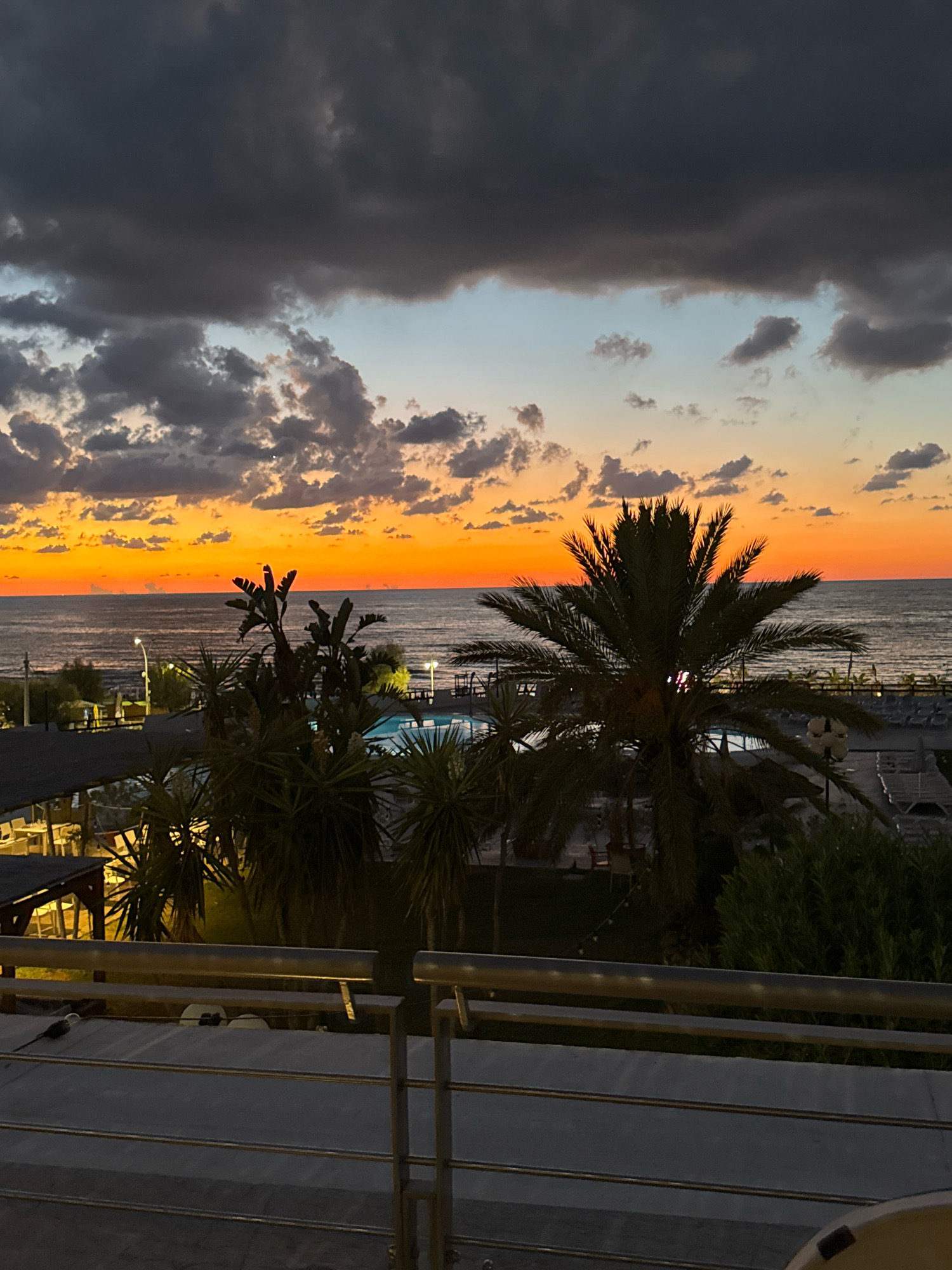 This is a photograph taken at dusk, looking out over the Mediterranean Sea from a vantage point in Isola delle Femmine, Italy. The sky is a dramatic display of color, transitioning from a deep, cloudy grey at the top to vibrant oranges and yellows along the horizon, reflecting on the calm water. Palm trees and other tropical vegetation are silhouetted against the sky in the foreground, partially obscuring a swimming pool and lounge chairs in the middle ground. Some buildings with awnings and outdoor seating are visible to the left, suggesting a resort or restaurant. A railing made of metal bars runs across the bottom of the frame, indicating the perspective is from a balcony or elevated walkway. There are no people visible in the image.