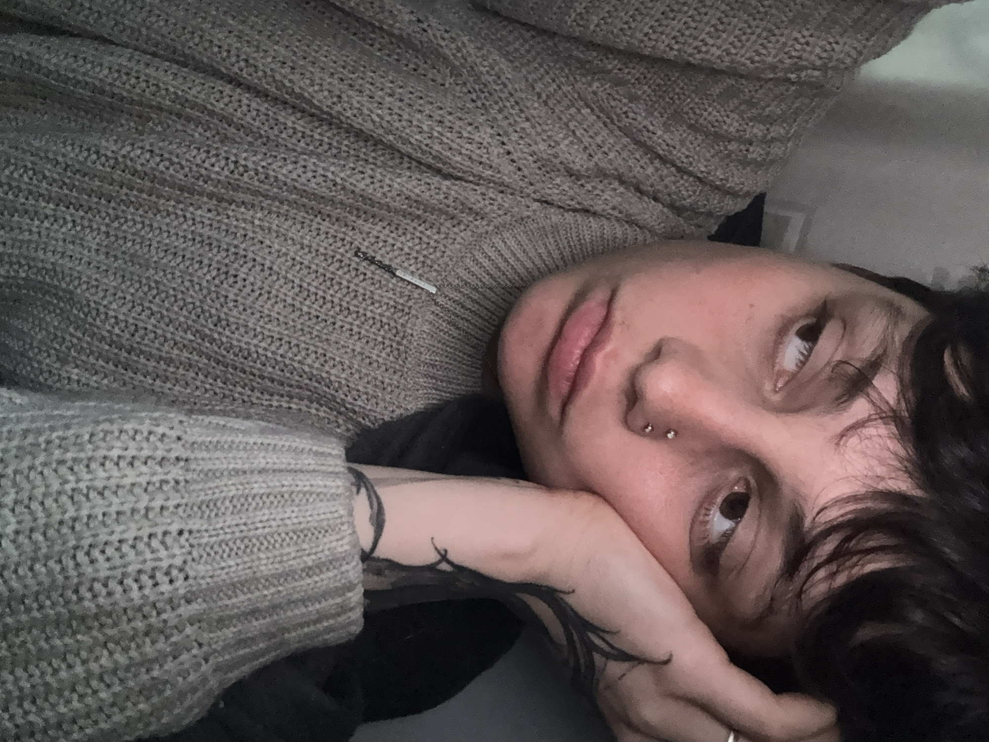 The image is a close-up portrait of a person lying down, viewed from a slightly high angle, with their head resting on their hand or arm. The person has dark, wavy hair, and is looking upwards and slightly to the right with a calm or pensive expression. They have distinct features including full lips and two small silver stud piercings on the bridge of their nose.
They are wearing a thick, light brown or grey knitted sweater, the texture of which is prominent across the upper part of the frame and on the visible sleeve. A thin silver necklace with a small, rectangular bar pendant lies across their chest; faint, indistinct text can be seen on the pendant, though it is not clearly legible. On their visible left forearm and hand, intricate black illustrative tattoos are visible, forming organic, flowing patterns that extend from beneath the sweater cuff. A simple ring is worn on one of their fingers.
The background appears to be a soft, light-colored surface, possibly a pillow or blanket, creating a cozy and intimate setting. The lighting is soft and ambient, suggesting an indoor environment. There are no visual cues within the image itself to indicate the provided location context of Saint Petersburg, Russia.