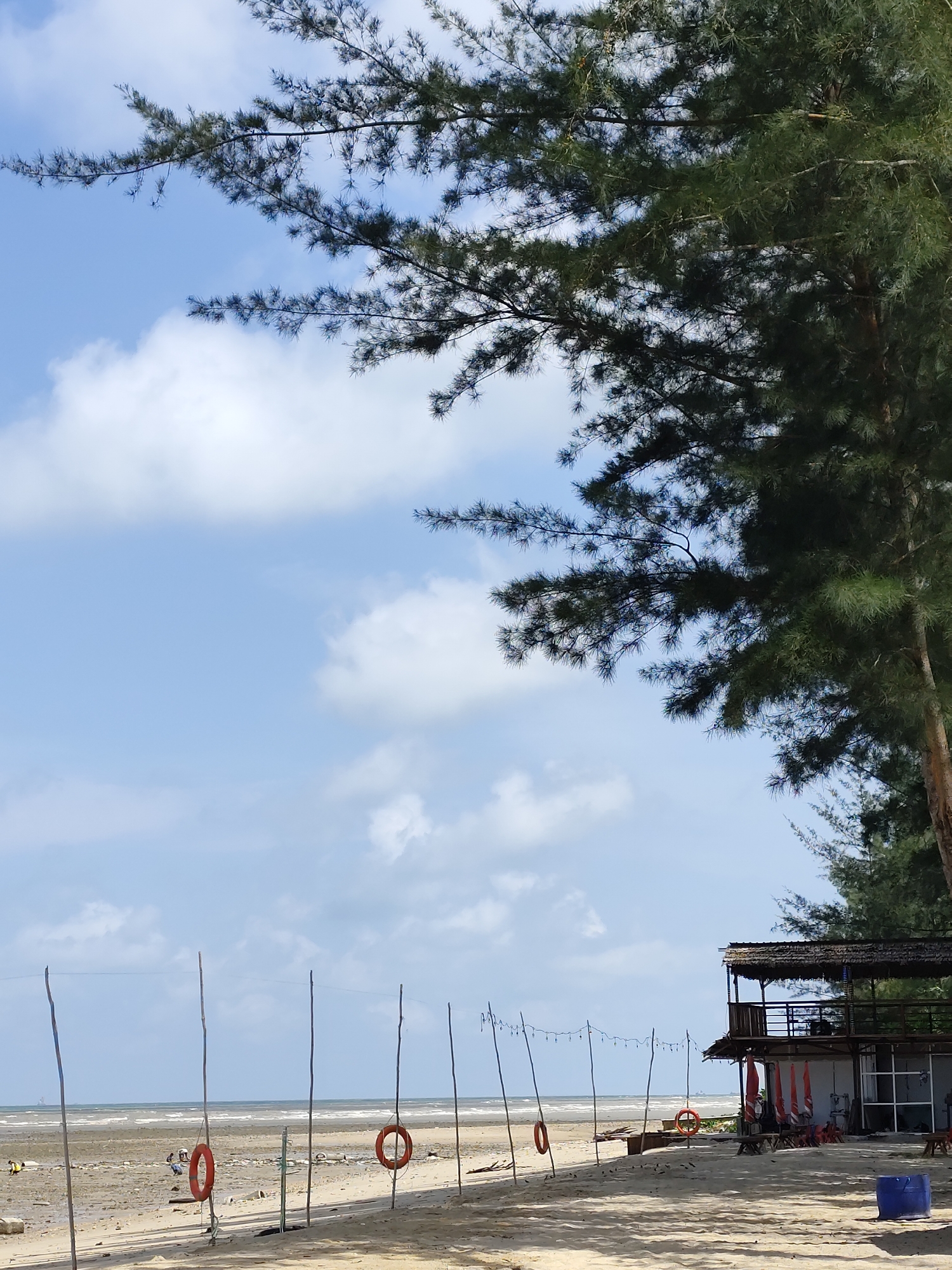 A sunlit, sandy beach with a calm ocean and a bright blue sky filled with scattered clouds. In the foreground, lush green tree branches cast shadows on the sand. Several wooden poles with orange life preservers attached are spaced along the shoreline. A two-story wooden structure, possibly a cafe or seating area, is visible on the right side of the image. There are a few scattered people on the beach in the distance, and some red umbrellas are set up near the structure. The tide appears to be out, revealing a wet, sandy shoreline.