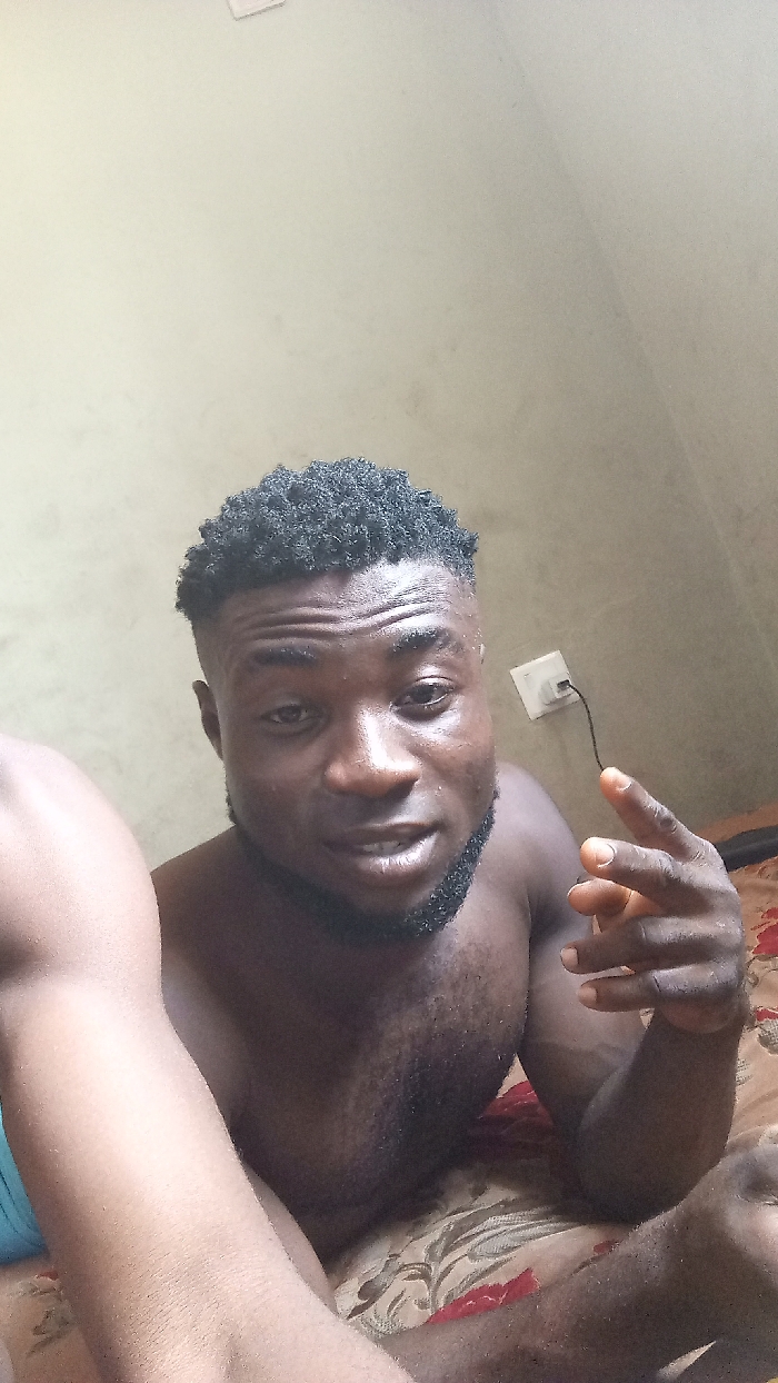 A shirtless, dark-skinned man with a short, textured haircut is shown in a selfie. He's partially reclined on a floral bedspread, with his arms visible. He has a dark beard. An electrical outlet with a connected cord is visible on the wall behind him. The wall is painted in a pale, neutral color. The overall lighting is somewhat dim.