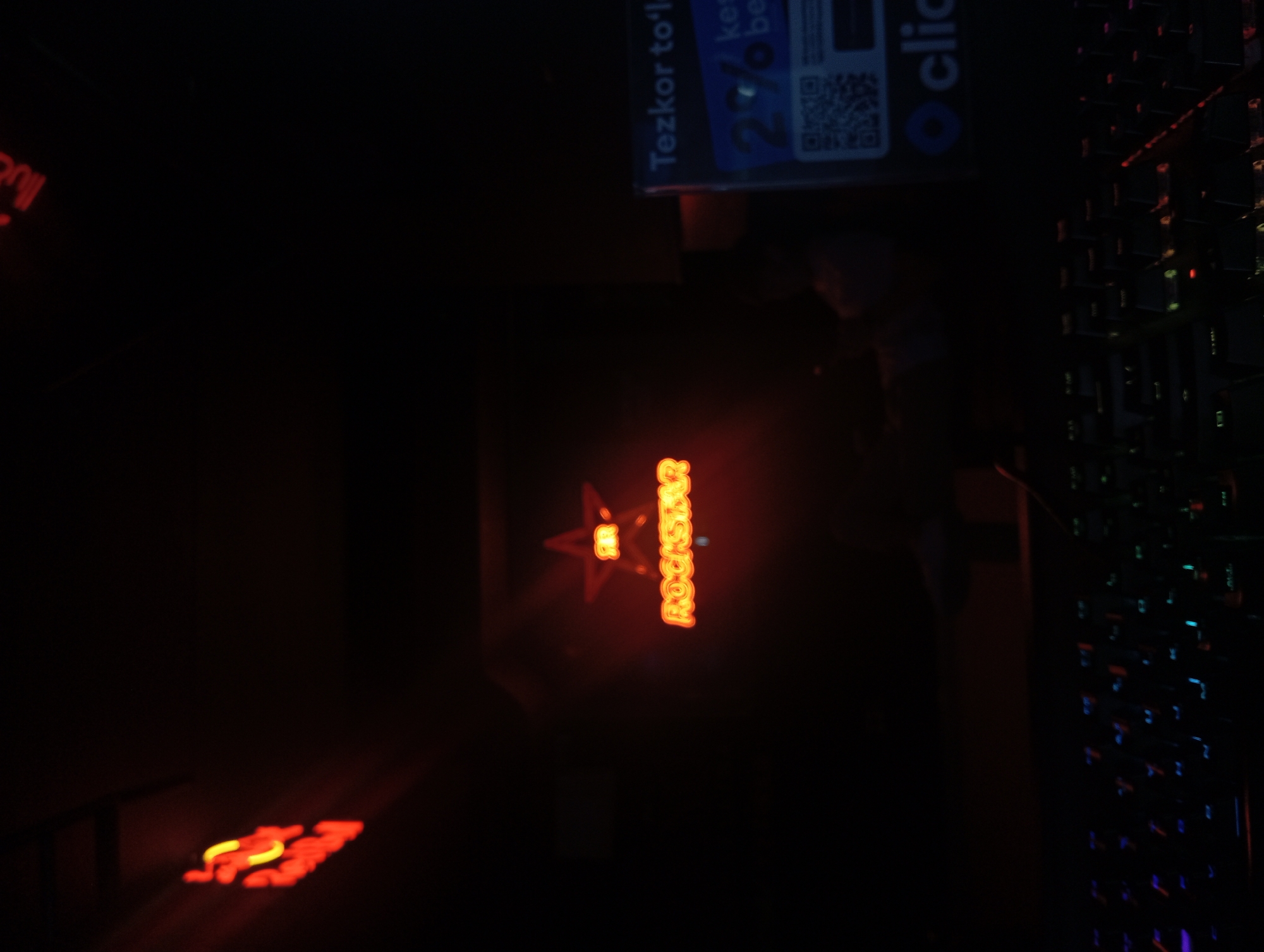 Here's a detailed description of the image:
**Scene:**
The image is taken indoors in what appears to be a dimly lit or dark environment. The overall lighting is poor.
**Objects:**
* A prominent "Rockstar" logo with a star above the word. The logo is illuminated in orange/red neon lighting.
* A keyboard with rainbow colored LEDs.
* A sign or advertisement for "Tezkor to'lov" and the logo of "Click".
* There is also what appears to be another sign, with an orange/red glow.
**People:**
* There appears to be a person or statue standing in the background.
**Location:**
Based on the text "Tezkor to'lov" which is in Uzbek, the image is most likely taken in Uzbekistan.
**Overall Impression:**
The scene gives the impression of a gaming setup or dimly lit space, with emphasis on the illuminated Rockstar logo and keyboard.