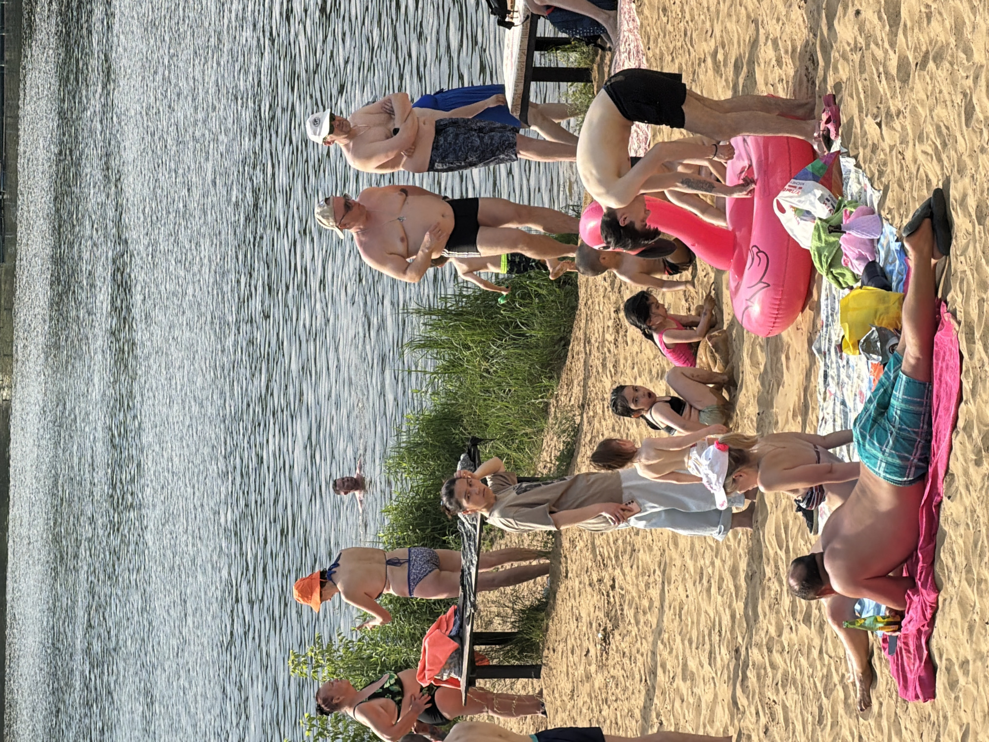 The image depicts a sunny day at a sandy beach on a lake in Malakhovka, Russia. Several individuals, including adults and children, are present. Some are in the water, while others are on the sand.
In the foreground, a man is lying on his back on a pink towel, facing away from the camera. He is wearing checkered green and blue shorts and has a bottle of beer next to him. A woman in a striped bikini is seated next to him.
Further back on the sand, a group of children are playing. A large, inflatable pink flamingo float is in the center of the image. Several towels and beach bags are scattered around.
In the middle ground, two women in swimwear are standing near a wooden table. One is wearing a green and black patterned bikini and the other an orange hat and blue bikini. Another woman in a tan t-shirt and light-colored pants is standing on the sand, looking towards the camera.
In the background, several men are standing near the water's edge, some with their arms crossed. A man is swimming in the lake. The lake is calm with ripples on the surface. Green foliage lines the edge of the water. The overall scene suggests a casual day of recreation.
