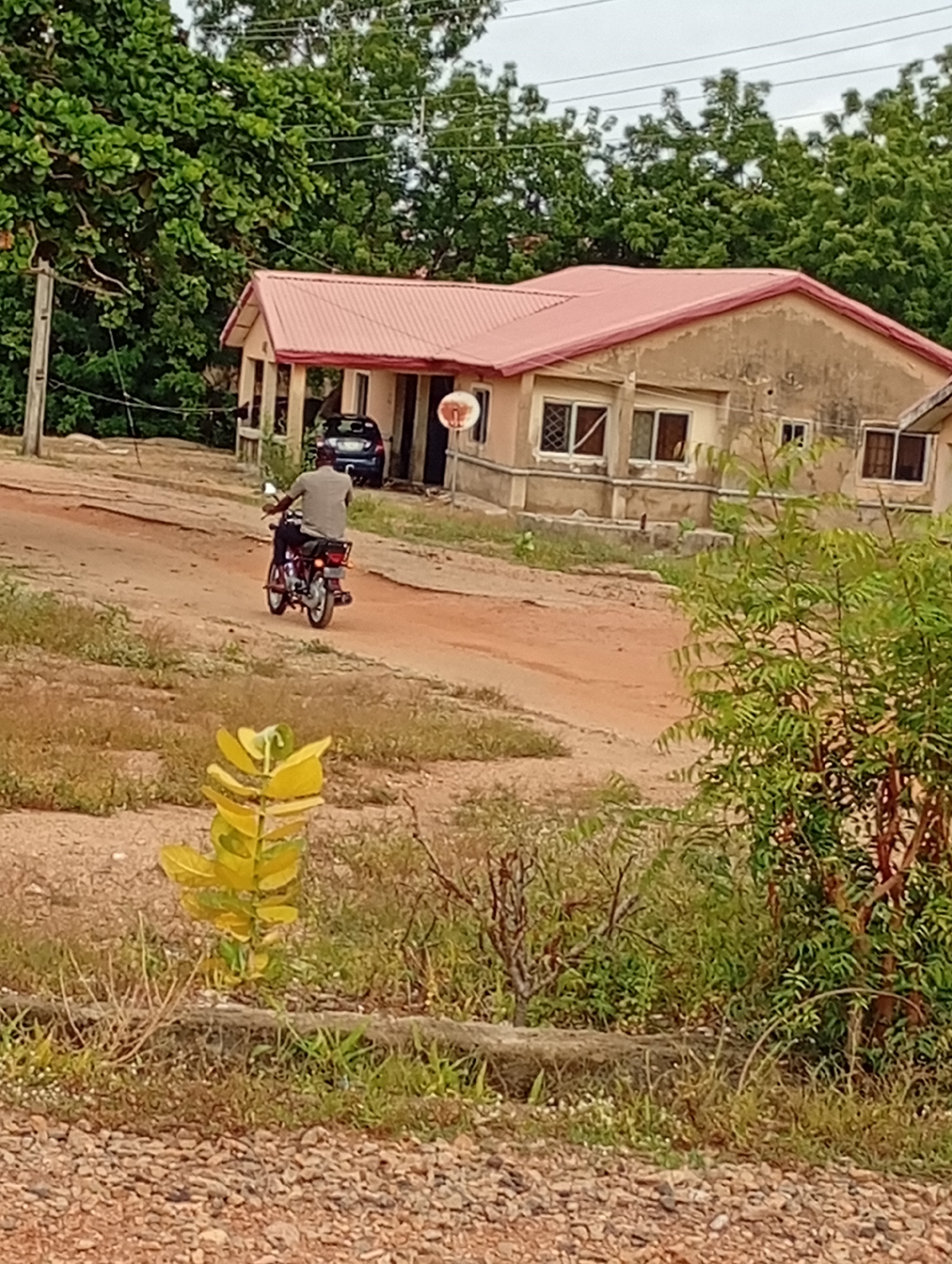 A man is riding a red motorcycle away from a house with a red roof in Adankolo, Nigeria. The house is beige with dark-framed windows. A blue car is parked in front of the house. The road is unpaved and reddish-brown. There are trees and shrubbery surrounding the house and road. A wooden utility pole is visible to the left of the house. The sky is overcast. A yellow plant is in the foreground.