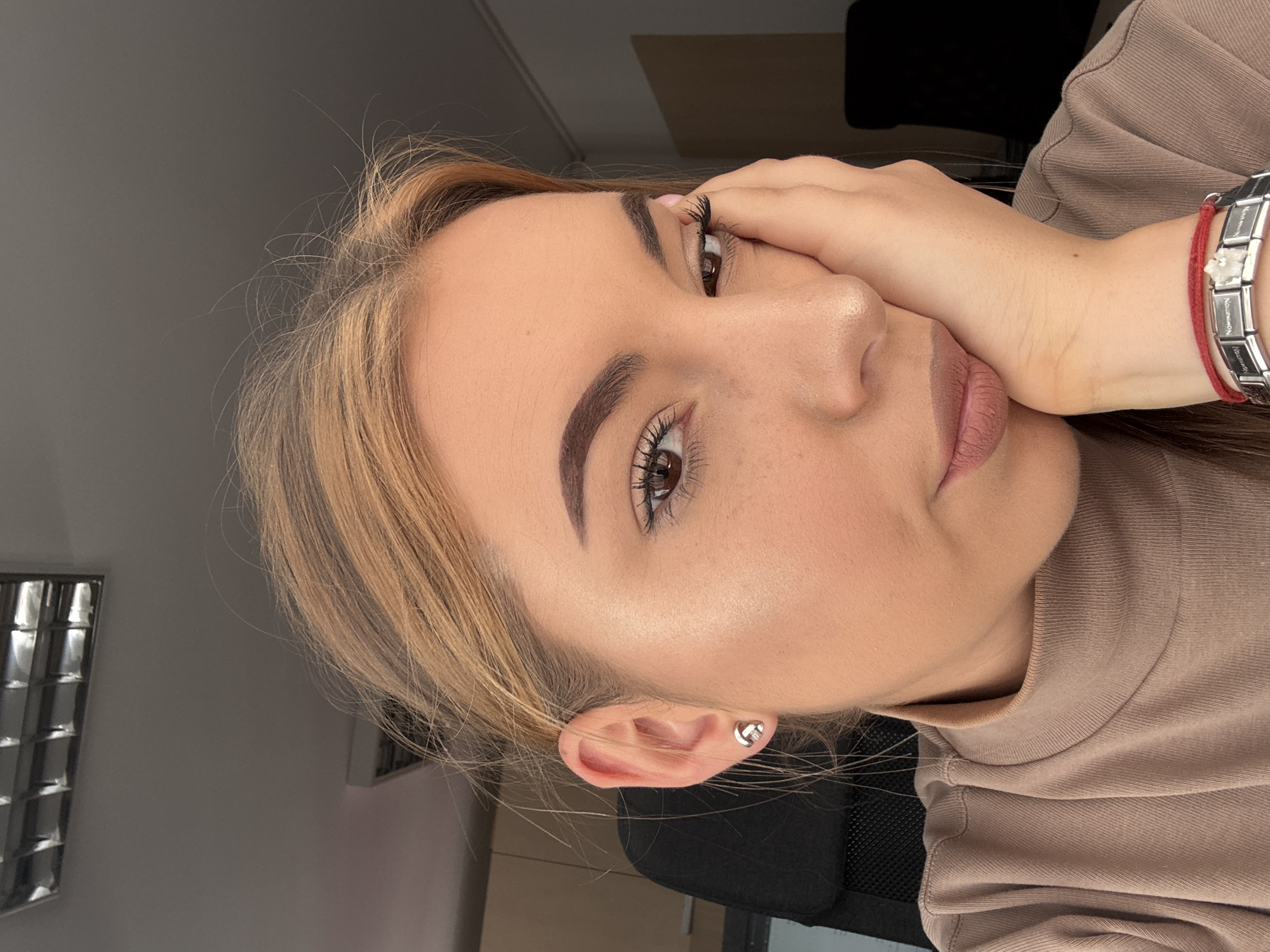 A young adult female, with light brown to blonde hair partially pulled back, a light complexion, and brown eyes, is depicted in a medium close-up. She wears a tan ribbed turtleneck top, a silver-toned stud earring with a knot-like design and a central small gem in her left ear, and multiple bracelets on her right wrist, including a red string bracelet and a silver-toned modular link bracelet by the brand Nomination, which features a white floral-shaped charm. Her makeup includes filled eyebrows, eyeliner, mascara, subtle highlight on her cheekbone, and a light pinkish-brown lip color. She rests her head on her right hand, looking towards the viewer with a thoughtful or slightly weary expression. Subtle freckles are visible on her nose and upper cheeks. The background indicates an indoor office environment with a plain grey wall and a standard rectangular recessed fluorescent light fixture on the ceiling. A black mesh office chair is partially visible behind her left shoulder. The scene is located in Sofia, Bulgaria.