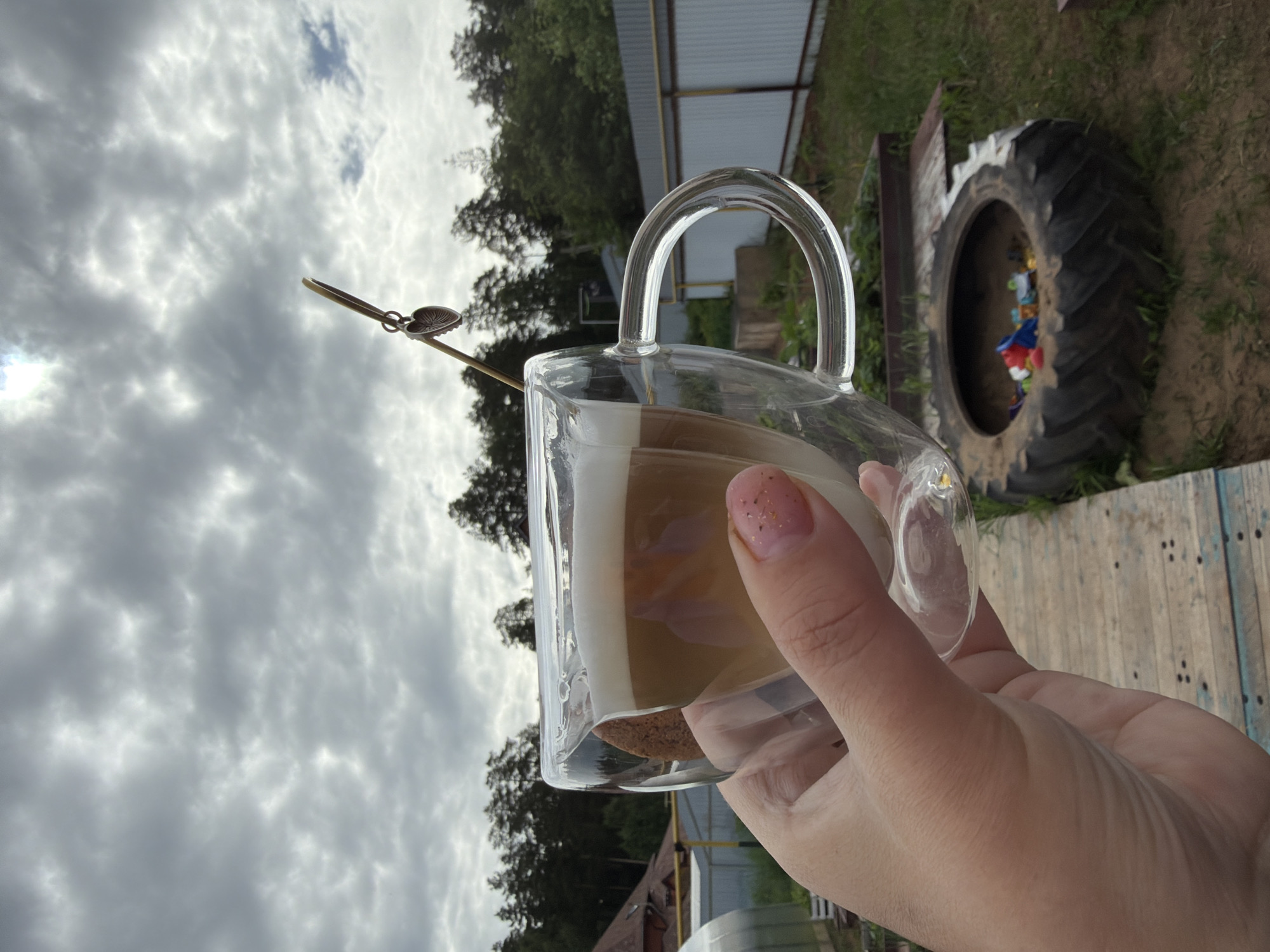 A close-up, eye-level outdoor photograph depicts a right hand holding a double-walled clear glass mug containing a layered beverage, likely a latte, against an overcast sky and a rural or suburban landscape. The top layer of the drink is white foam, followed by a light brown liquid, and a darker brown liquid at the bottom. A round, light brown cookie is visible between the inner and outer walls of the mug, partially submerged in the beverage. A thin, gold-colored stirring stick with a small, metallic heart-shaped charm is inserted into the drink, angled upwards towards the right. The person's thumb and index finger are visible, with the index finger having light pink nail polish with small glitter specks.
The background shows a cloudy sky, with some areas appearing brighter as if sunlight is attempting to break through. Below the sky, a dense line of green coniferous trees extends across the horizon. To the left, a portion of a house with a dark roof and a greenhouse with a clear, curved roof are visible. A yellow pipe runs horizontally in the mid-ground. To the right, a gray corrugated metal fence separates the property from another area. A wooden pathway or decking with blue and weathered planks is visible in the lower left foreground. In the lower right foreground, a large black tire is repurposed, likely as a planter or sandbox, containing various colorful plastic items resembling children's toys. The ground is a mix of dirt and patchy green grass. The scene is located in the city of Chaikovsky, Russia.