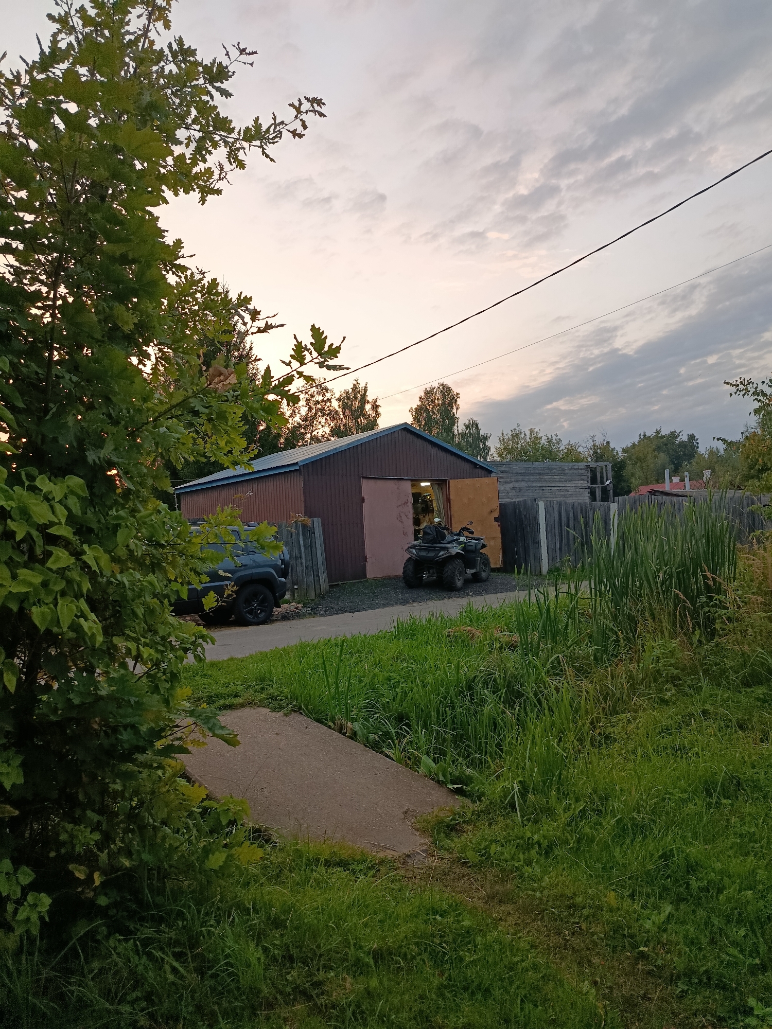 In the city of Vereya, Russia, an outdoor scene unfolds near a brown metal-sided building with a wide, open doorway. A dark SUV is parked to the left of the building's entrance. Inside the open doorway, an ATV with a person on it is visible. A weathered wooden fence runs along the right side of the image, behind which are tall reeds and other vegetation. The ground in front of the building is covered with gravel. A concrete path or sidewalk leads from the foreground towards the building. Lush green grass and foliage dominate the foreground and right side of the image, with a large tree with green leaves on the left. The sky above is overcast with a soft, diffused light, suggesting either early morning or late evening. A power line stretches across the upper portion of the frame.