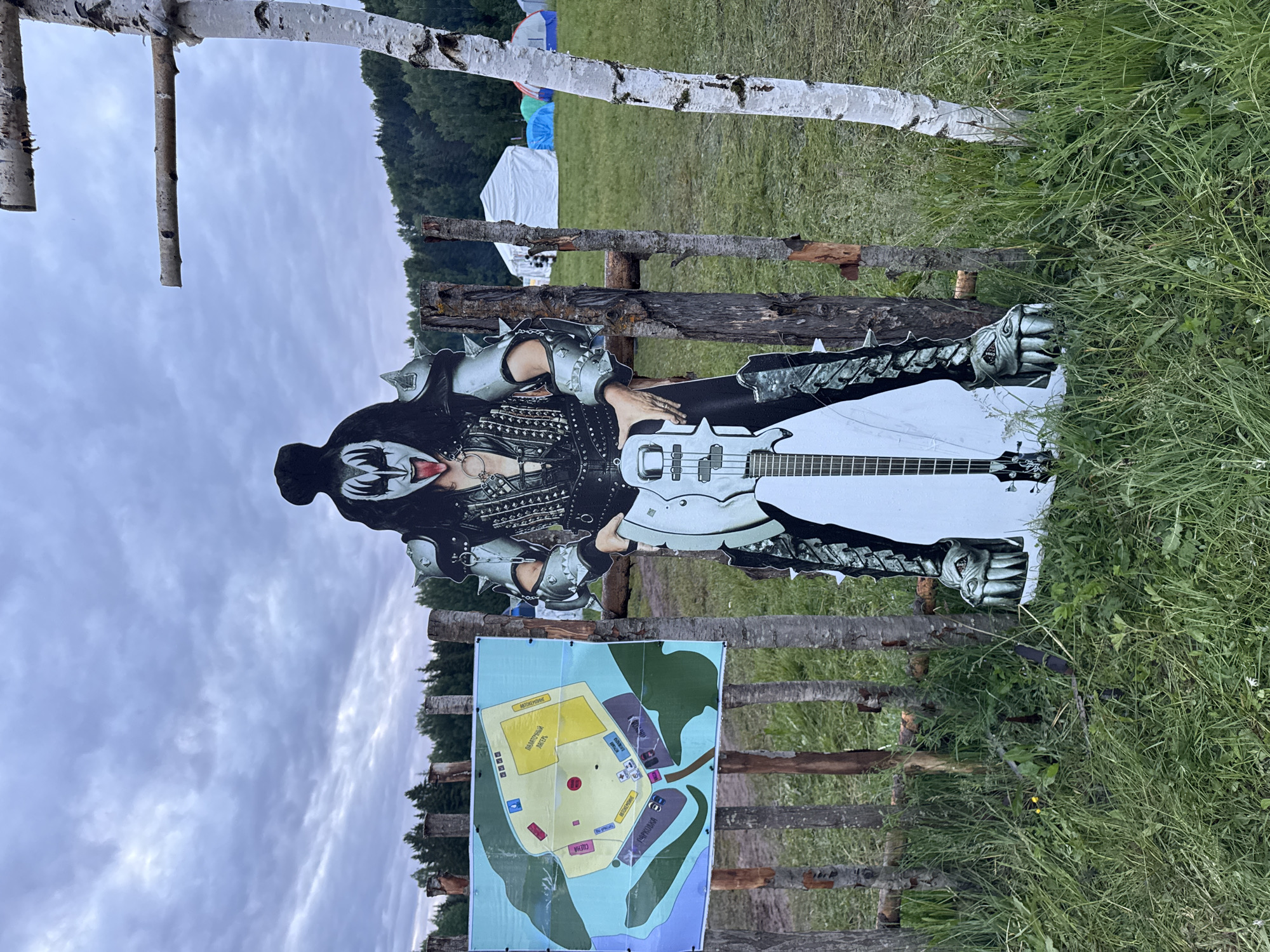 The scene, set in Большая Кивара, Russia, captures an outdoor area under an overcast sky. A rustic fence constructed from vertical and horizontal rough-hewn wooden logs or branches delineates the foreground. Attached to the left side of this fence is a laminated map displaying a layout plan with various colored sections and Cyrillic text, indicating areas such as "ПАЛАТОЧНЫЙ ЛАГЕРЬ" (Tent Camp) and "ПАРКОВКА" (Parking). To the right of the map, directly behind the fence, stands a life-sized cardboard cutout of Gene Simmons from KISS, in his "Demon" persona, featuring black and white facial makeup, a spiky silver and black costume, and his tongue extended. He holds a white bass guitar. The ground is covered with green grass and scattered weeds. In the background, beyond the fence and cutout, an open grassy field extends to a distant forest line. A large white event tent is visible in the mid-ground, alongside several smaller camping tents in blue and green. On the far right, a white-barked tree trunk with two short, horizontal branches stands vertically. The lighting is soft and diffused due to the uniformly cloudy sky.