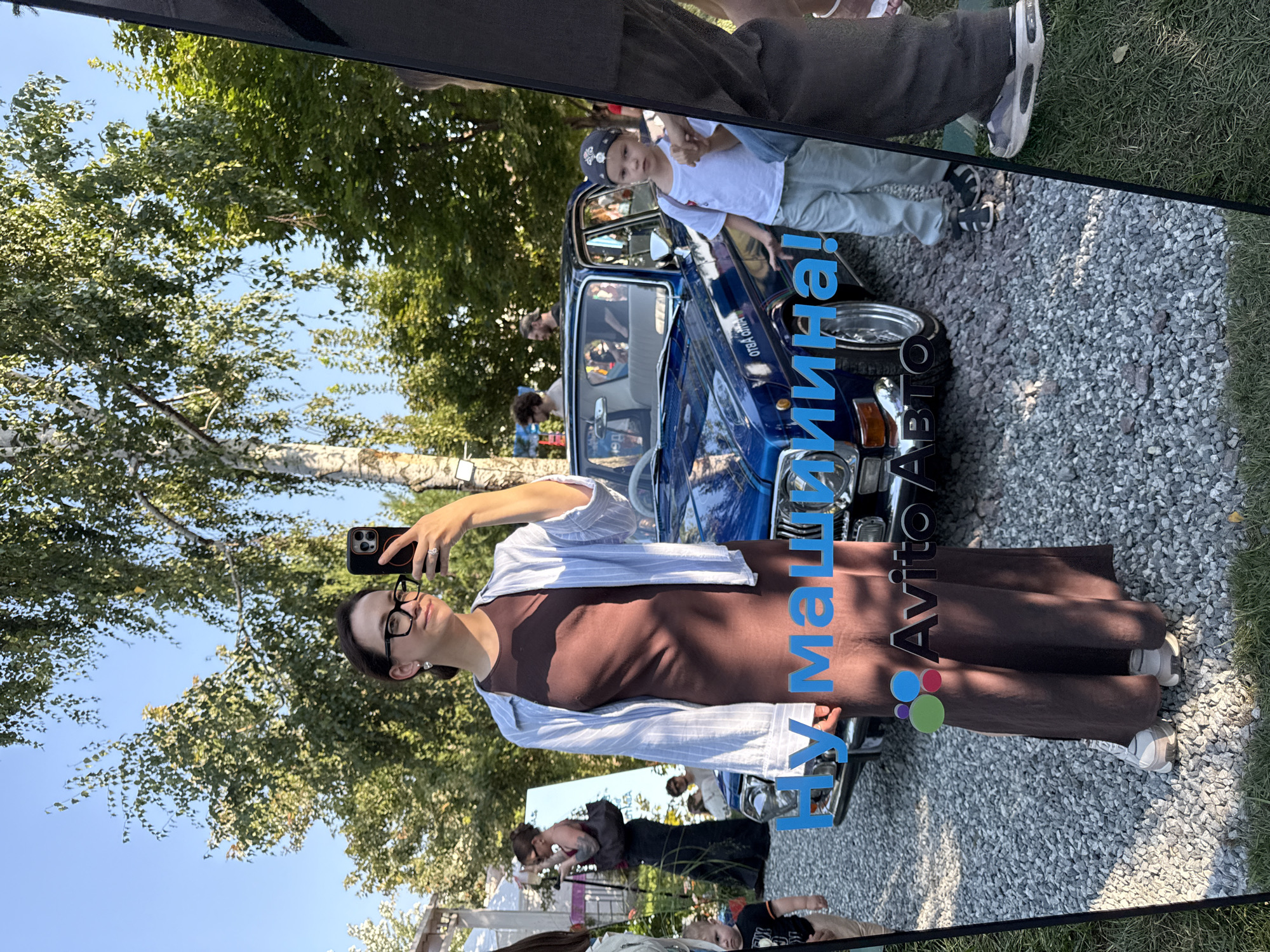 The image is a selfie taken by a woman with glasses and a brown dress, holding a phone. She is standing in front of a blue car, likely at an outdoor event or car show. The reflection in a mirror shows other people and elements of the environment, including trees and a blue sky. The reflection also displays text that appears to be in Russian. The text "Ну машина!" translates to "Well, the car!" and "Avito Авто" is likely a reference to a Russian classifieds website for cars. Without additional context or visible landmarks, it is impossible to definitively determine the location within Kazan, Russia.