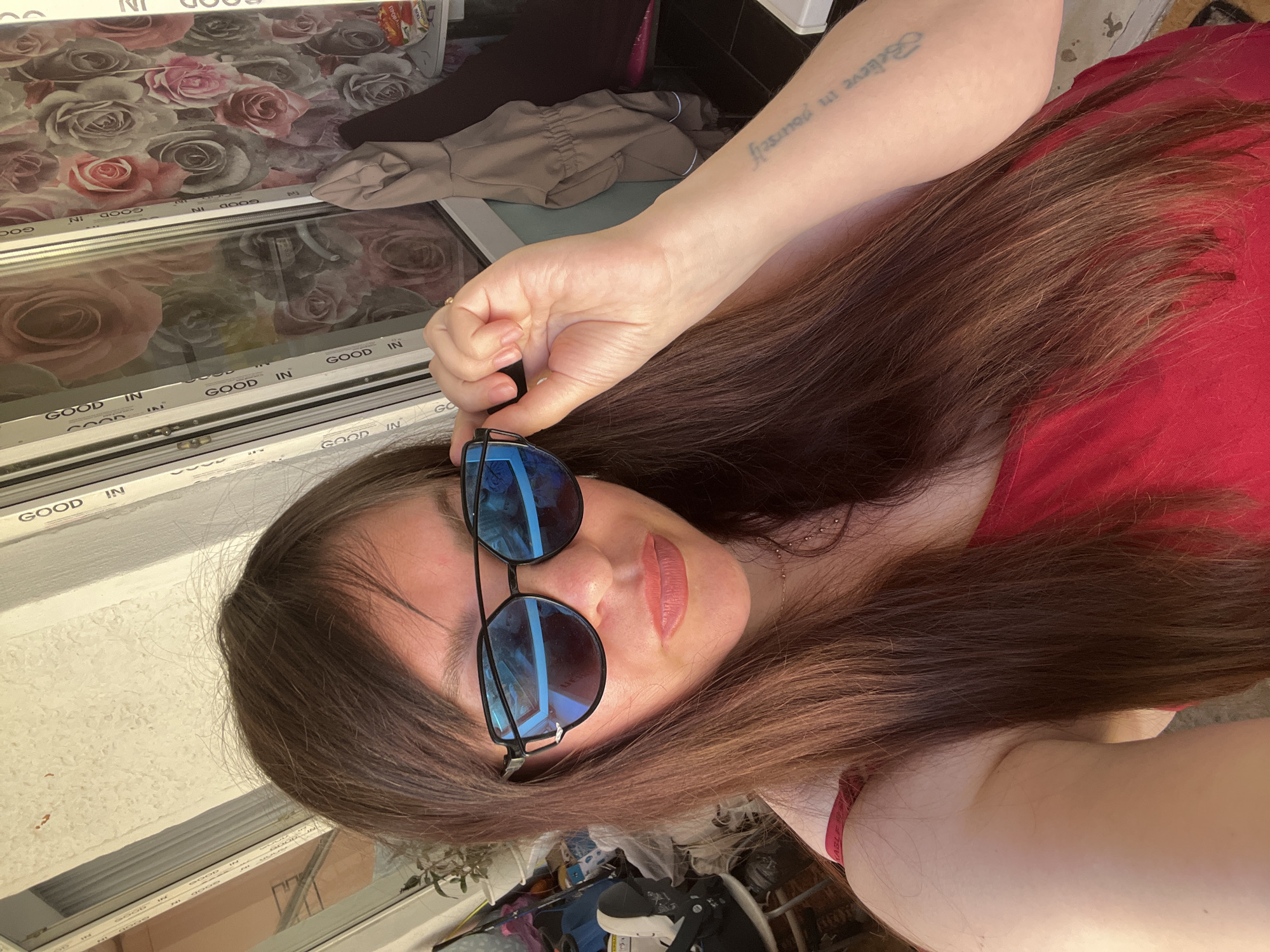 A woman with long, brown hair, wearing a red top and sunglasses, is captured in a close-up, self-taken photograph. She is holding the arm of her sunglasses with her right hand, and her left arm is resting at her side. A tattoo is visible on her left forearm. The background suggests an outdoor setting with a window that has a floral wallpaper pattern, and what appears to be household clutter in the lower left corner of the frame. The overall lighting indicates it is daytime, with sunlight illuminating the scene.