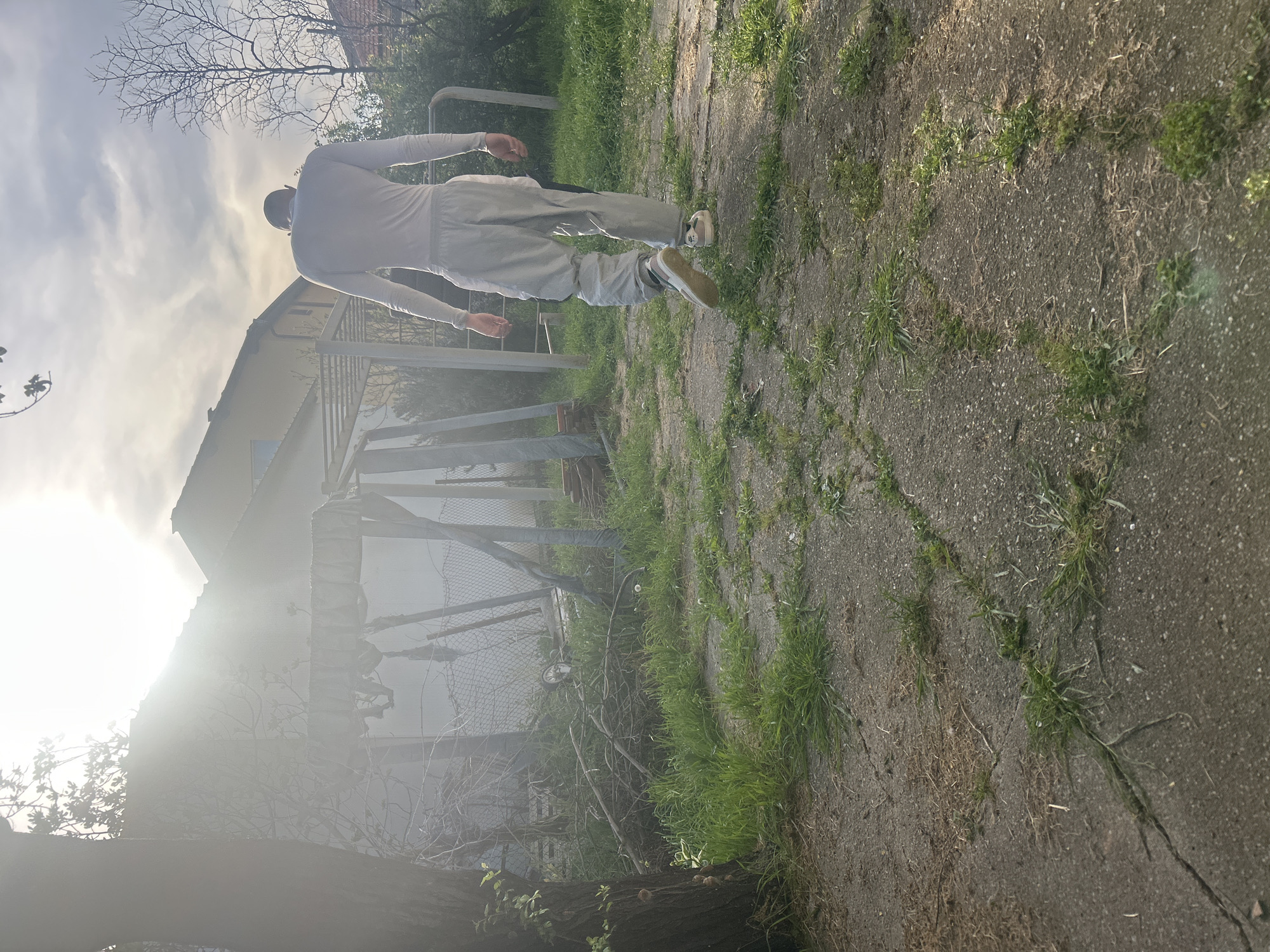 A person in light-colored clothing is walking away from the viewer on a cracked concrete path interspersed with patches of grass. The scene appears to be in a backyard or garden area. A metal fence and some dilapidated structures are visible in the background, partially obscured by a tree and bright sunlight. The location is possibly in Burgas, Bulgaria. The sky is overcast with bright sunlight breaking through.