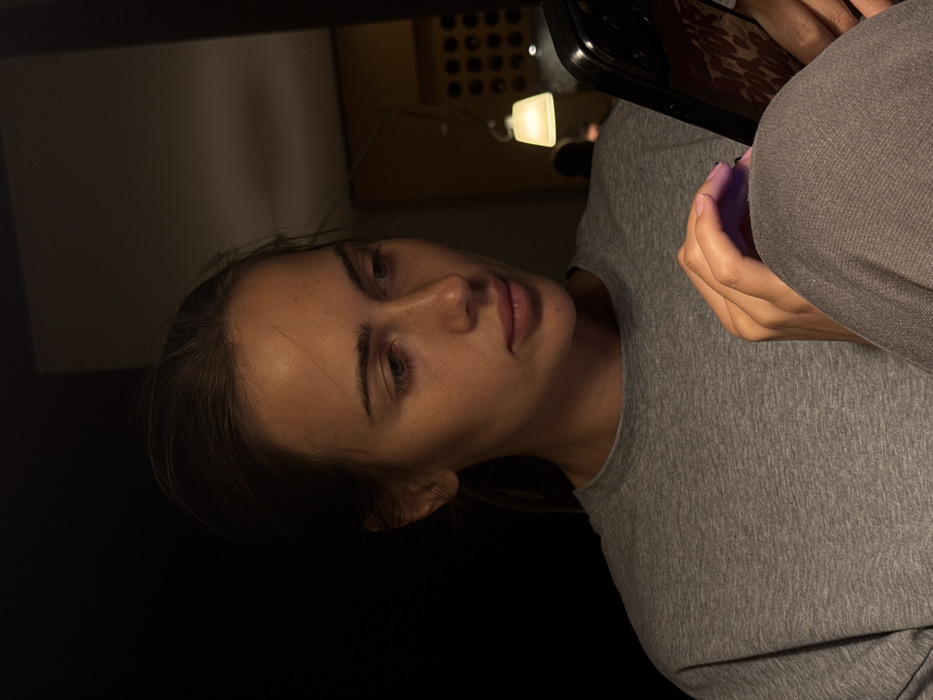 The image displays a young adult female positioned in a dimly lit indoor environment. She possesses light skin, dark hair (pulled back), and visible freckles across her nose and cheeks. Her attire consists of a grey crew-neck t-shirt and grey pants. Her gaze is directed downward, focused on a dark-cased smartphone held in her left hand, while her right index finger appears to be interacting with the device's screen. The smartphone case features a reddish-brown pattern. Warm artificial light illuminates her face and hands. In the blurred background on the right side of the frame, a hanging light fixture with a translucent cream-colored shade emits a soft glow. Behind this fixture, a dark wall or structure with multiple circular indentations arranged in a grid pattern is visible. A faint, out-of-focus silhouette of another individual is partially discernible behind the light. The specific location within the city of Warsaw, Poland, cannot be determined from the visual information present in the image.