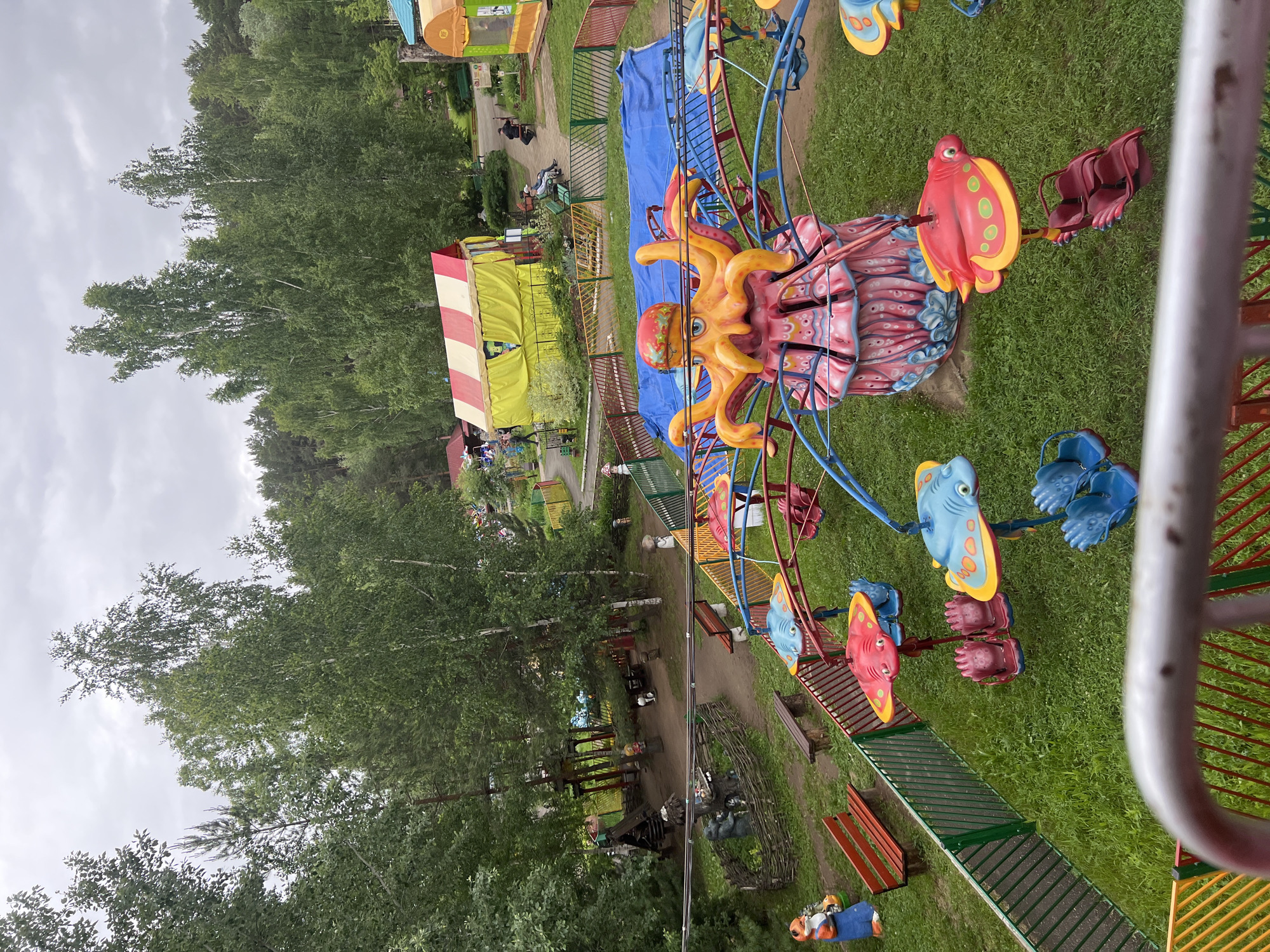 This image presents an outdoor amusement park or children's recreation area located in the city of Chaikovsky, Russia, under an overcast sky. In the immediate foreground, a silver metal railing partially frames a colorful, circular children's ride. This ride features a central pillar designed as an orange octopus with a red cap, supported by a pink and blue base, from which several brightly colored vehicles shaped like red and blue stingrays or fish are suspended, each containing two empty seats.
The midground landscape includes patches of green grass, dirt pathways, and various decorative elements. To the left, a blue fox statue stands near a red and orange wooden bench. A green and red metal fence divides the ride area from a path leading to a rustic wooden fence enclosing two brown bear statues. Additional small statues, including mushrooms and gnomes, are visible throughout the grounds. Further back, a large yellow and red striped tent structure is prominent, with a smaller yellow and green tent situated to its right. Two individuals are seated on a green bench to the right of the striped tent, and a third person is seated near the yellow and green tent. The background consists of a dense forest composed of both deciduous and coniferous trees, beneath a uniformly gray, cloudy sky. No active use of the ride or specific interactions among the visible individuals are discernible.