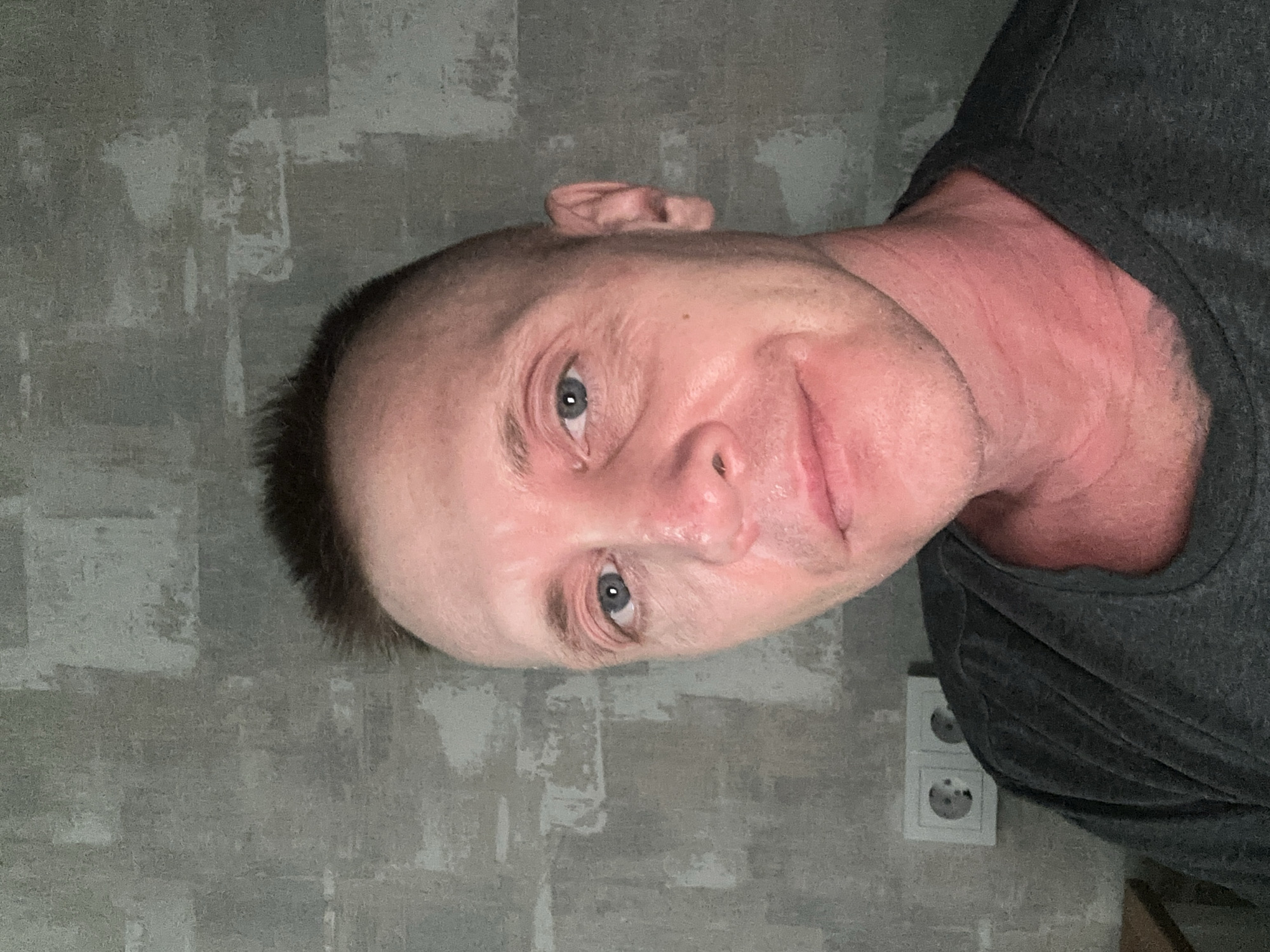 A close-up, angled portrait of a fair-skinned man with a shaved head and blue eyes. He is wearing a dark gray shirt. The background is a gray wall with textured, abstract patterns. A double European-style electrical outlet is visible on the wall behind him. The image was likely taken indoors. There is no definitive evidence the photo was taken in Oryol, Russia.