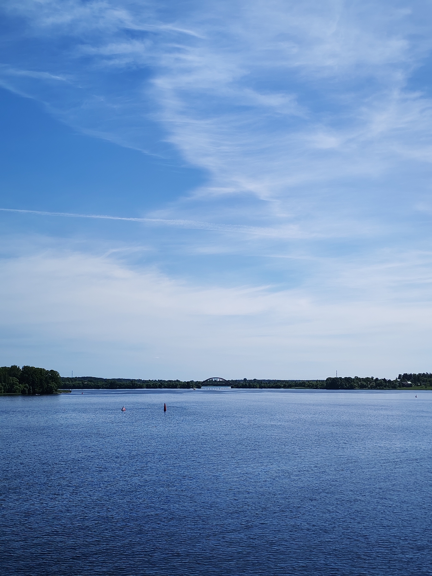 The image displays a wide landscape view of an expansive body of water under a vast sky, with a distant, forested horizon. The sky occupies approximately the upper two-thirds of the frame, showing a clear light blue hue with scattered white, wispy cirrus clouds. A faint, straight white contrail is visible horizontally across the upper mid-section of the sky. The lower third of the image is filled with dark blue water, exhibiting subtle ripples across its surface. Two buoys are present in the water: a conical red buoy in the mid-ground, slightly right of center, and a less distinct, lighter-colored cylindrical buoy positioned further back, to the left of the red buoy. The horizon line reveals a narrow band of dense green trees and foliage, indicative of a distant shoreline. Above this treeline, a slender utility pylon is visible slightly left of center. To the right, a dark, arched bridge spans the water in the distance. The scene is consistent with the landscape of the Volga River, characteristic of the area near Kalyazin, Russia. No people or active vessels are visible.