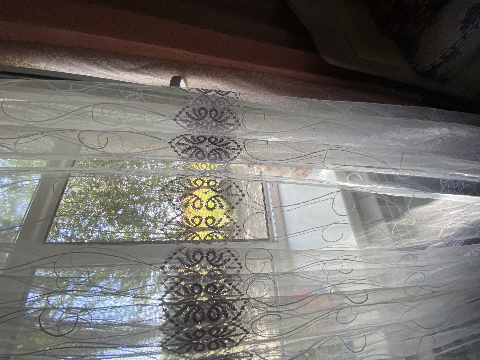 The image captures a view through a sheer white curtain with embroidered detailing. The curtain has a repetitive floral pattern in a dark grey or purple thread, forming a horizontal band across the fabric. Above this band, the curtain features swirling, thread-like patterns. Through the curtain, a window reveals a bright outdoor scene with lush green trees and foliage illuminated by sunlight. The light creates a dappled effect on the outdoor scene. To the right of the sheer curtain, a heavier, darker curtain, possibly reddish-brown or rust-colored, is partially visible, drawn to the side. The overall impression is of an indoor setting looking out onto a bright, natural environment. The location is identified as Keŋ-Say, Kyrgyzstan.