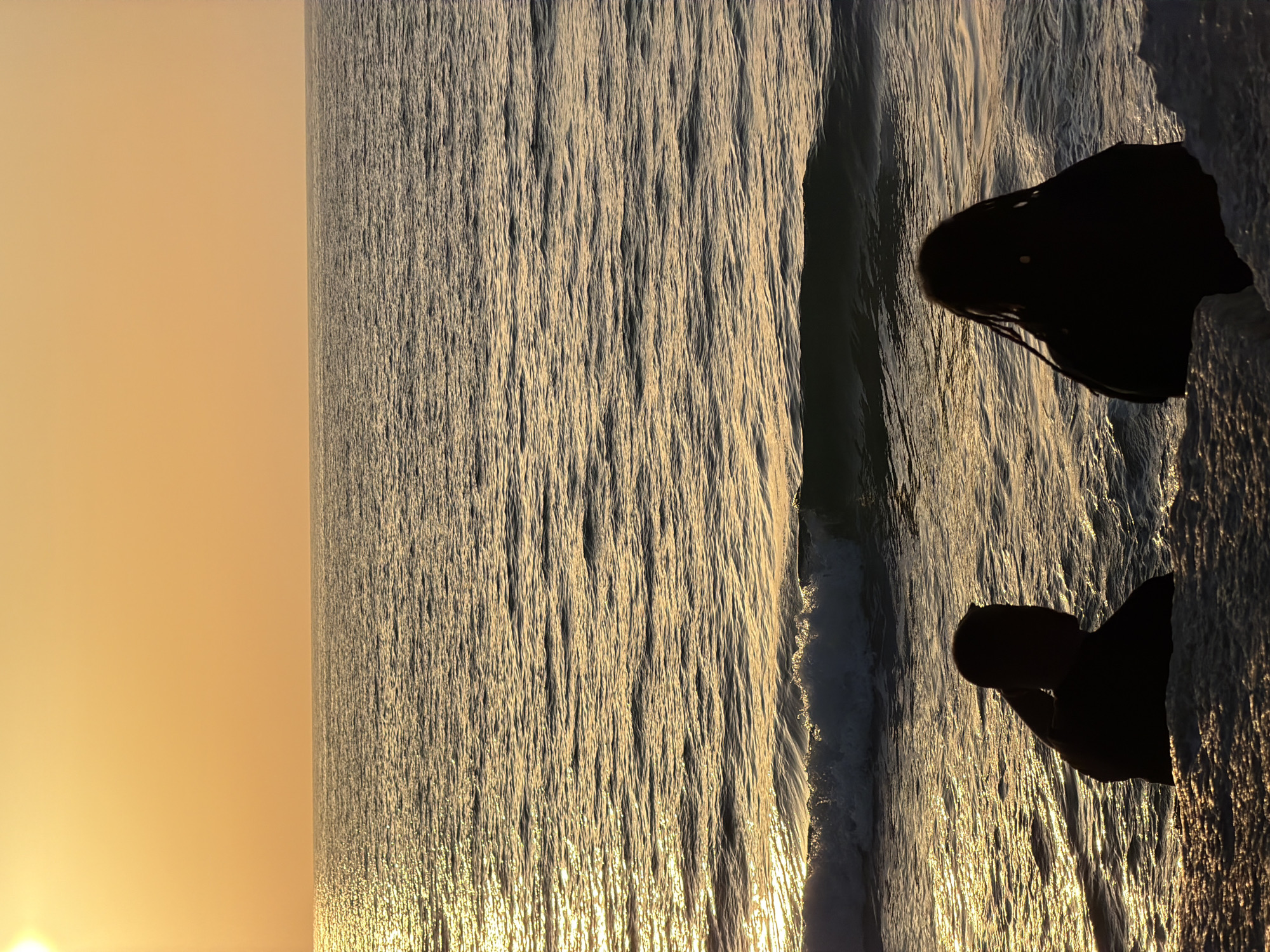 The image depicts a sunset scene on the Mediterranean Sea in Tel Aviv, Israel. The sun is low on the horizon, casting a golden light on the water and sky. The sea appears textured with small waves reflecting the sunlight. The silhouettes of two individuals are visible on a stone structure at the edge of the water. One figure has long hair.