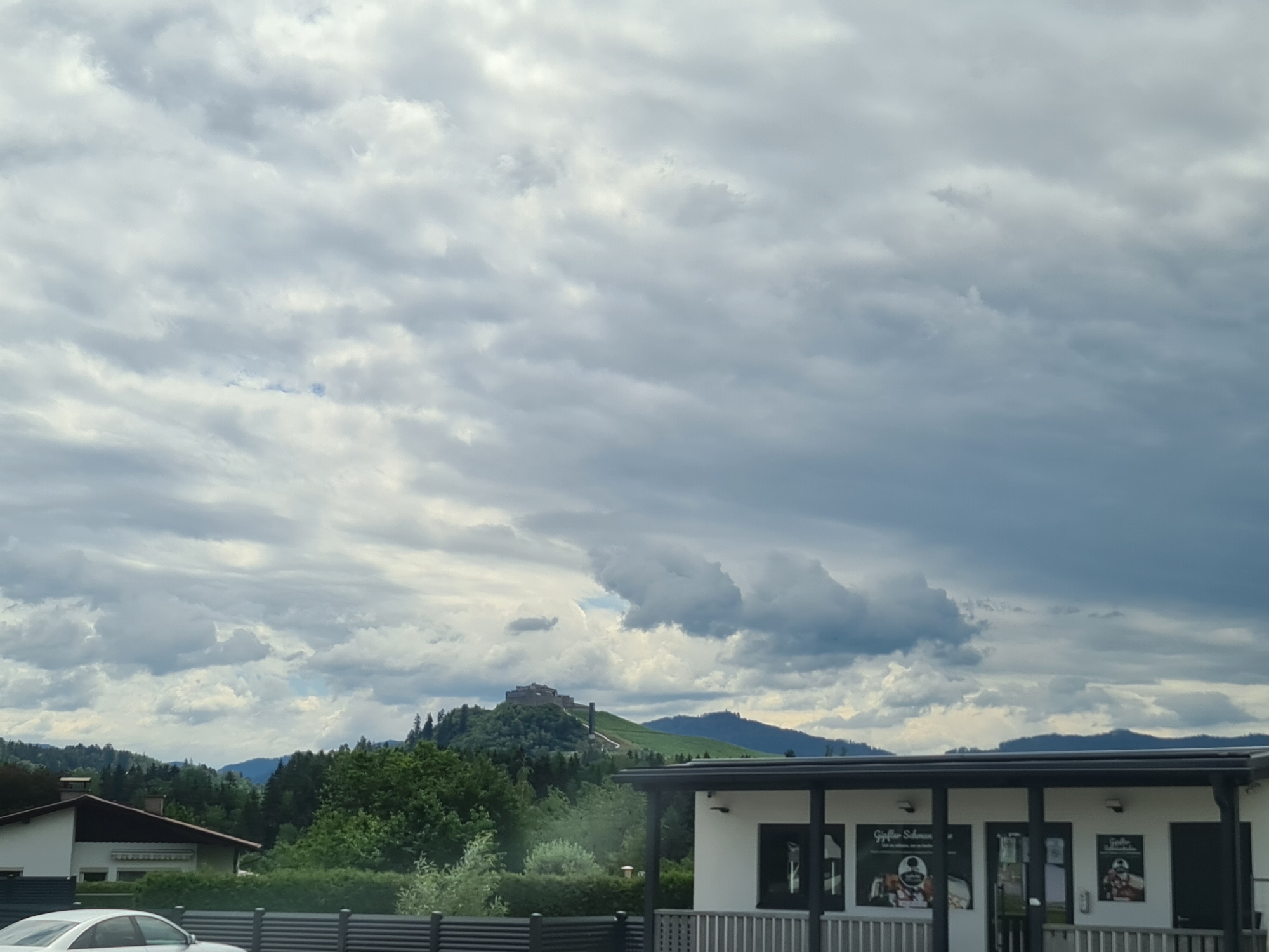 The image depicts a cloudy sky over a landscape with a building in the foreground and a castle on a hill in the background. The building in the foreground is a modern, single-story structure with a black roof and white walls. It appears to be a commercial establishment, as indicated by signage on the windows. The building has a covered porch area supported by black pillars. In front of the building, a black fence runs horizontally across the frame. To the left of this building, a portion of a house is visible, with a white wall, a dark roof, and a chimney. A white car is parked in front of the fence.
In the middle ground, a densely wooded hill rises, covered in green trees. The castle is situated atop this hill, a prominent structure with stone walls and towers, appearing somewhat ruined but still substantial. A path or road is visible winding up the side of the hill towards the castle.
The background consists of rolling hills and mountains, covered in trees, fading into the distance under a heavy, overcast sky with large, dramatic clouds. The overall scene suggests a rural or semi-rural setting in or near the city of Frauenstein, Austria, given the presence of the castle, a common feature of the Austrian landscape. The lighting is diffused due to the cloudy conditions.