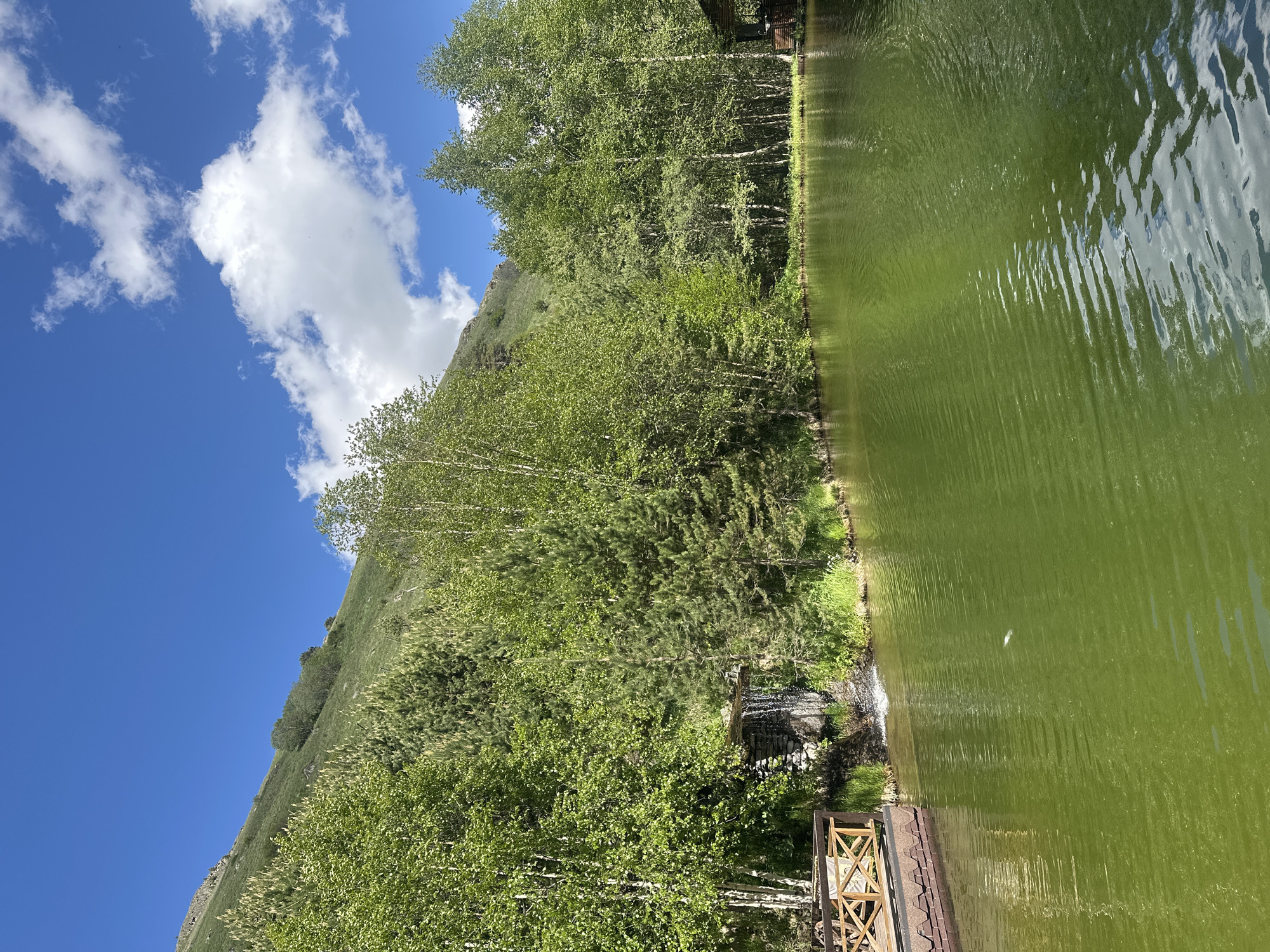 The image shows a serene lake with green water, reflecting the blue sky and white clouds. A small waterfall cascades into the lake from a rocky structure surrounded by lush green trees. On the left side of the lake, a wooden deck with a railing is visible, extending over the water. The background is dominated by a steep, grassy mountain slope covered in trees, typical of the natural landscape around Terskol, Russia. The scene is tranquil and idyllic, suggesting a natural resort or park setting in the Elbrus region.