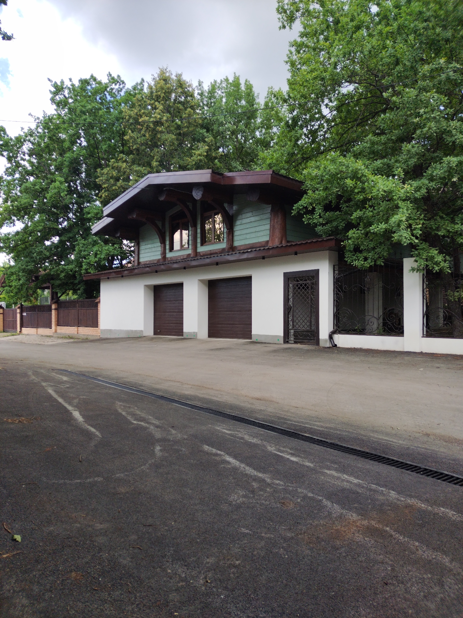 The image depicts a two-story house with a distinctive architectural style, likely a garage attached to the main residence. The lower level features two dark brown garage doors, set within a clean white facade. Above the garage, the second story is clad in a pale green material and features a steeply pitched, dark brown roof with exposed wooden beams, creating a rustic yet modern aesthetic. Two large windows on this upper level reflect the surrounding trees and cloudy sky.
The house is situated behind a white fence and a decorative wrought-iron gate, hinting at a private property. To the left, a wooden fence with brick pillars marks the boundary of the property. Mature trees with lush green foliage frame the house, suggesting a setting with abundant nature. A paved driveway with a drainage grate runs in the foreground.
The scene appears to be set in the daytime, under an overcast sky with some clouds visible. The lighting suggests it could be late afternoon or early evening. There are no people visible in the image, and the overall mood is quiet and serene, with no clear activity taking place. The location context of Blokhino, Russia, places this scene in a possibly suburban or rural setting within the country. There is no visible text in the image.