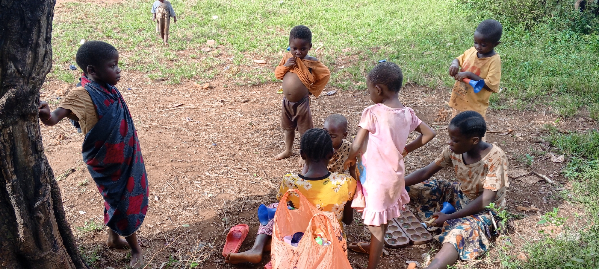 This outdoor image captures a group of approximately eight children and young people gathered in Ngalula, Democratic Republic of the Congo, under bright daylight. The scene unfolds on reddish-brown dirt, with patches of green grass and taller vegetation in the background. A prominent, dark tree trunk borders the left side of the frame.
The children are engaged in various casual activities. On the far left, a young person in a light-colored shirt and a red and blue patterned wrap leans against the tree, looking to the right. In the middle, a young boy stands barefoot, with his orange shirt pulled up, exposing his bare stomach, and wearing brown shorts. Another child in the background wears a light grey t-shirt with "NINJA TURTLES" faintly visible and walks away from the camera.
In the foreground, several children are seated on the ground. One young person wears a yellow floral top, with two orange plastic bags beside them. Another child, wearing a light brown patterned top, is seated, looking downwards, potentially engaged in a game. A traditional Mancala-style board game, containing seeds or stones, lies on the ground near these children, suggesting play. Standing nearby, with their back to the camera, is a child in a pink dress, and another child in an orange "VE SPORT" t-shirt holds a blue funnel-like object. Most children are barefoot, and their expressions are generally neutral or curious. The setting implies a relaxed, everyday social or play gathering in a rural or semi-rural area.
