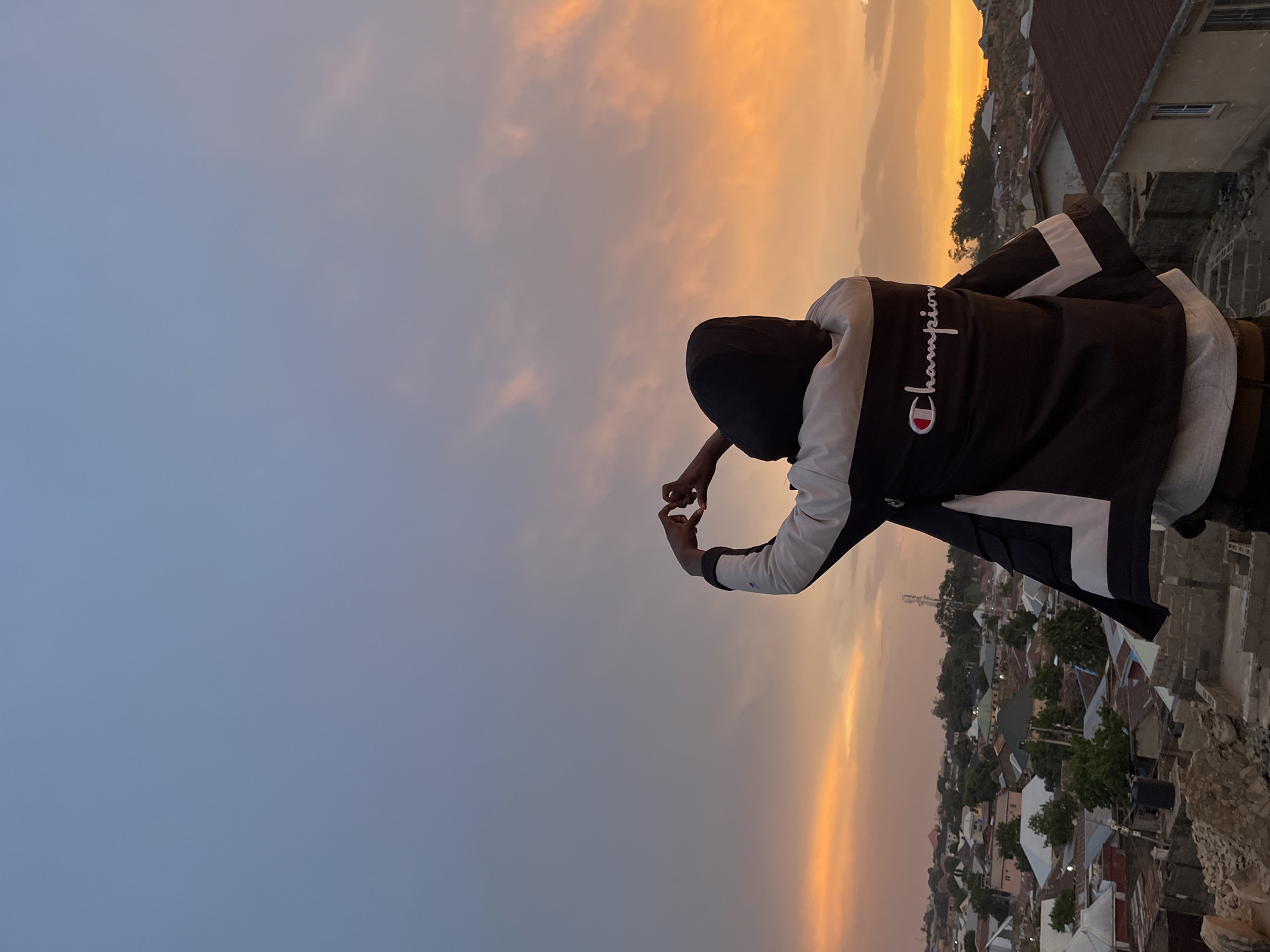 A person stands on a rooftop at dusk in Jos, Nigeria, making a heart shape with their hands against a vibrant sky transitioning from blue to orange near the horizon. The person wears a dark jacket with white trim and the word "champion" written in white with the company's logo on the back, and a dark hoodie obscuring their face. Below, a densely populated cityscape is visible with buildings of various heights and roofing materials. A cell tower is partially visible in the background. The image is rotated 90 degrees counter-clockwise.