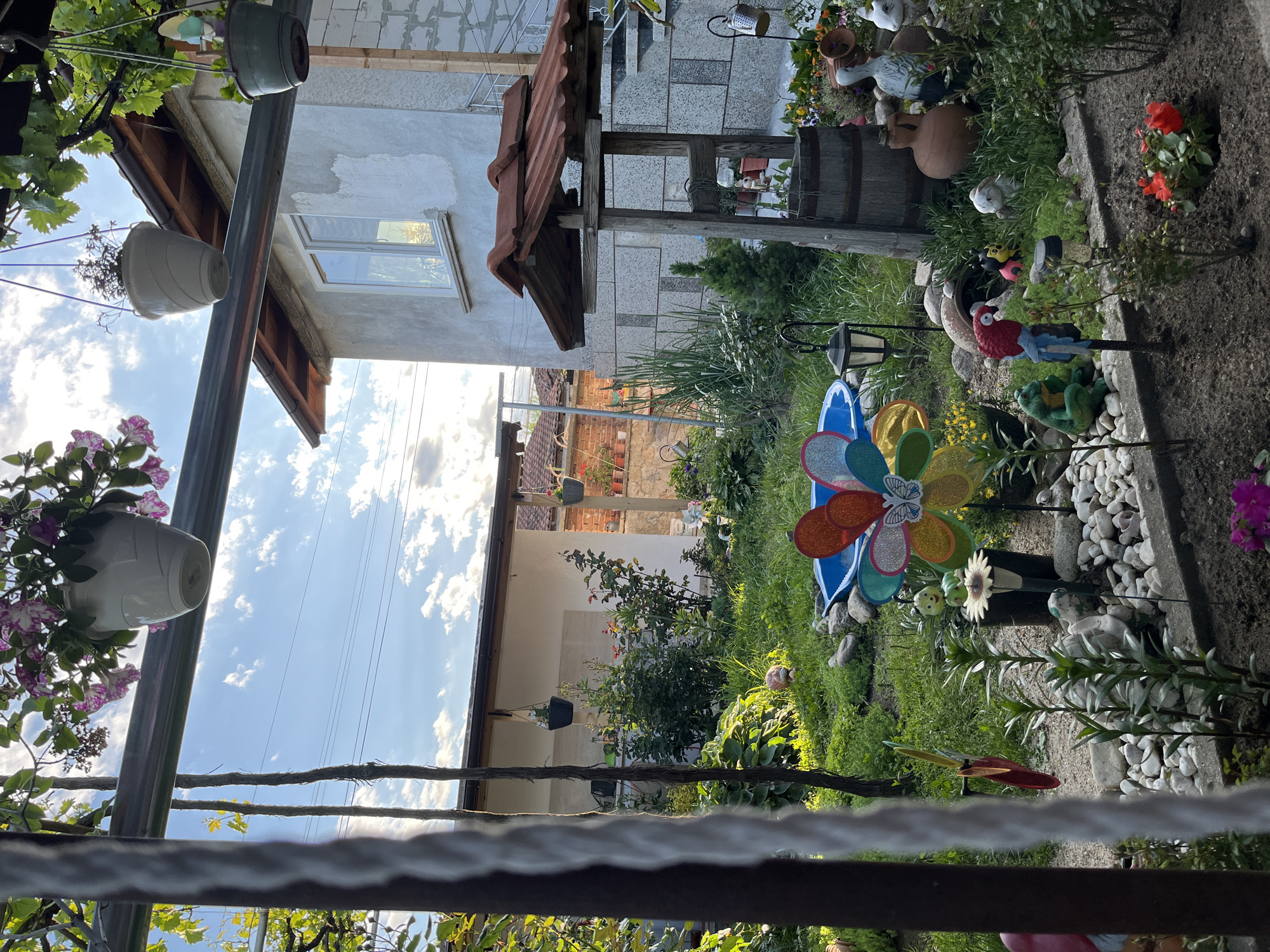 The image depicts a backyard garden scene in Karavelovo, Bulgaria, viewed from a partially obstructed perspective. A variety of garden decorations are present, including pinwheels, flower pots, and ceramic figures amongst foliage and white gravel paths. The architecture visible includes portions of residences with tiled roofs and plastered walls. Overhead, a pergola structure with hanging planters is partially in view. The sky is blue with scattered clouds.