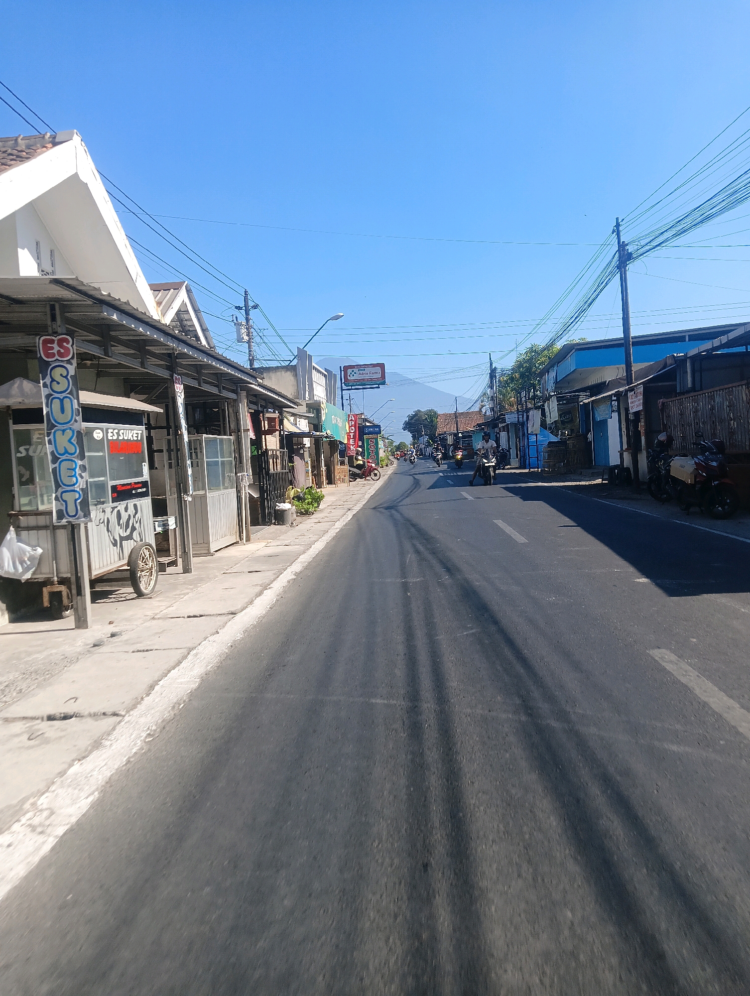 The image captures a street scene in Sokaraja, Indonesia, under a clear blue sky. The perspective is from a moving vehicle, likely a motorcycle, looking down a paved road lined with shops and stalls on the left side.
On the left, a food stall selling "ES SUKET" (grass jelly ice) is prominently featured, with its signage clearly visible. Adjacent to it are other small businesses, some with open fronts and display cases. Utility poles with numerous wires run along the street, a common sight in Indonesian towns.
The road itself shows tire marks, suggesting regular traffic. Several motorcycles are visible on the road, with some individuals riding them. In the distance, mountains are faintly visible against the horizon. The overall impression is of a typical day in a moderately busy street in a Southeast Asian town.