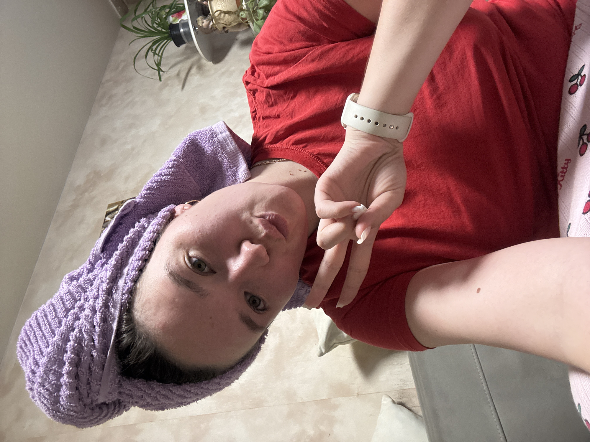 A selfie features a person with fair skin and brown hair partially wrapped in a light purple towel. They are wearing a red T-shirt and pink pajama pants with cherry patterns. The person is making a peace sign with their right hand. A white smart watch is on their left wrist. In the background, there is a neutral-colored wall and a potted plant. The location appears to be indoors. There is no indication of Фоки, Russia, within the image.