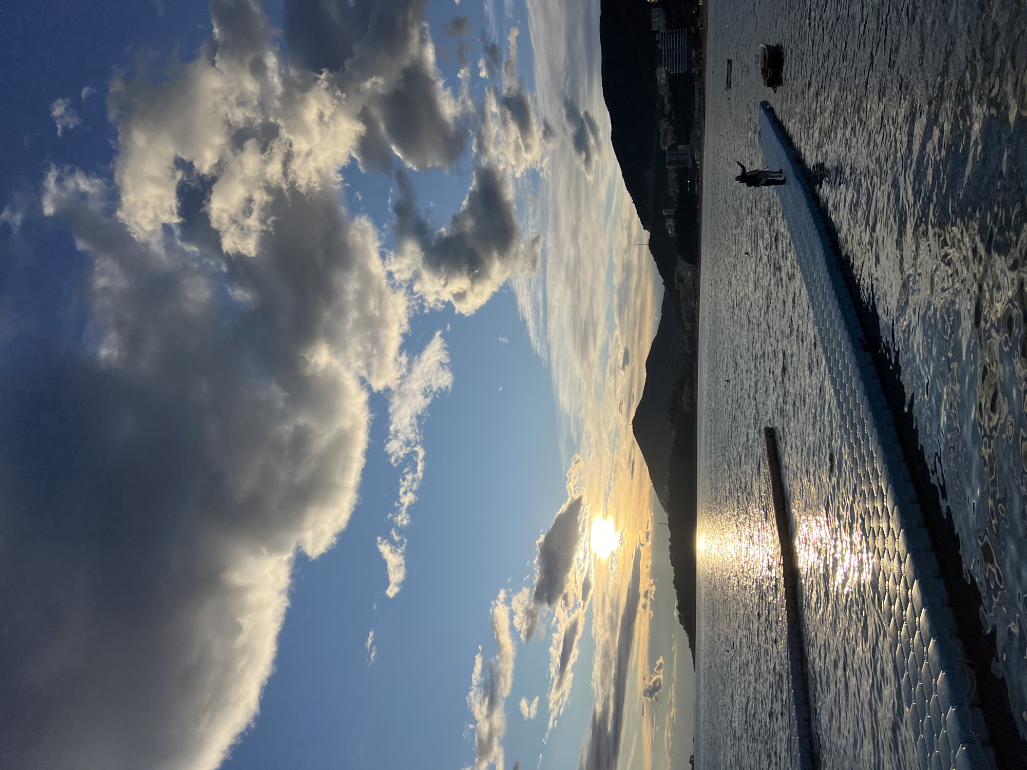 The photograph captures a scene at sunset over the water in Budva, Montenegro. The sky is filled with white and gray clouds, with the sun partially obscured by the mountains in the background. A long, narrow pier extends into the water, with a person standing at the end, seemingly observing the sunset. The water reflects the sunlight, creating a shimmering effect. Buildings are visible along the coastline in the background.