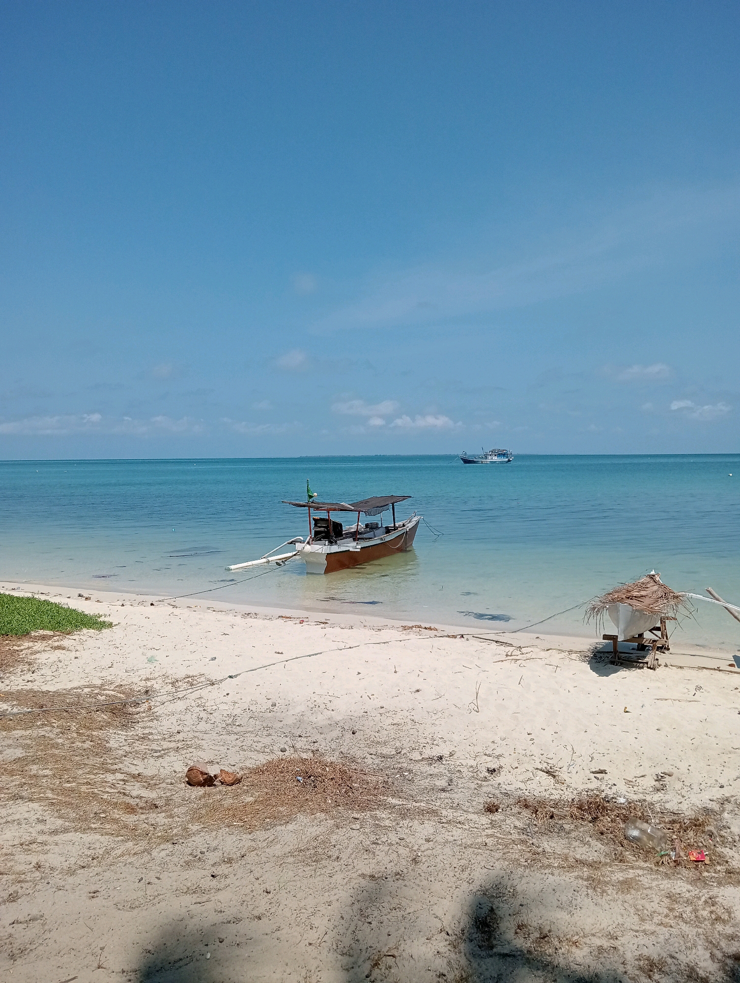 The image depicts a tranquil beach scene in Pagerungan Kecil, Indonesia, under a clear blue sky with a few scattered clouds. The foreground is a sandy beach with scattered dried foliage and a coconut. To the left, a patch of green vegetation meets the sand. In the shallow, turquoise water near the shore, a brown and white fishing boat with a canopy is moored. Further out in the sea, a larger boat is visible on the horizon. To the right of the foreground boat, a small, rustic shelter constructed from palm fronds sits atop a wooden frame, with a boat hull partially visible beneath it. The water is calm and clear, revealing the sandy bottom in the shallows.