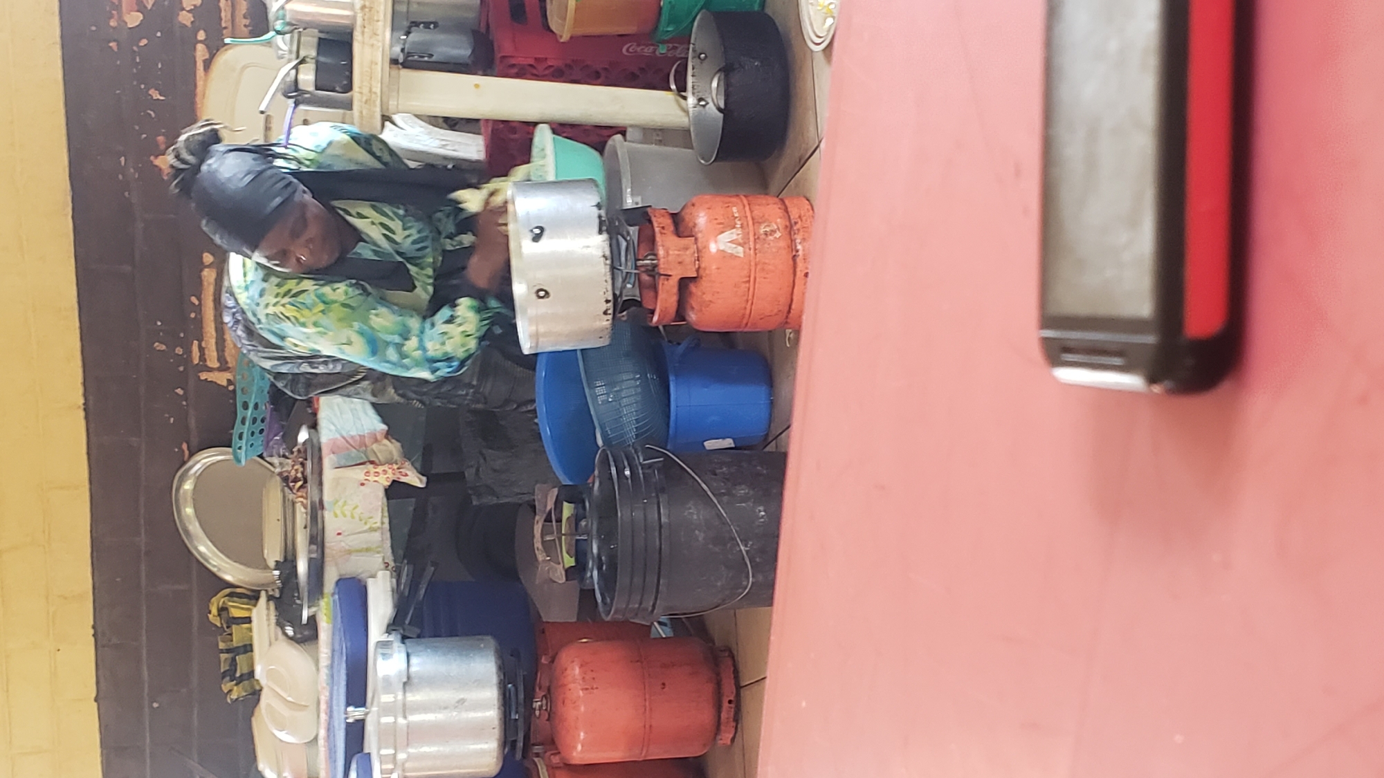 In Unguwar Shanu, Nigeria, a woman wearing a headscarf and a patterned long-sleeved shirt is engaged in cooking outdoors. She is crouched beside a portable gas stove with a pot on top, appearing to be washing or cleaning something. Surrounding her is a collection of kitchenware and cooking supplies, including large metal pots, plastic buckets of various sizes and colors (black, blue), and a blue colander. Two orange gas cylinders are prominently visible, one directly beneath a pot and another to the left. The setting appears to be an open-air or semi-enclosed area, possibly a market or an outdoor kitchen, with a brick wall and a light-colored ceiling overhead. In the foreground, a pink surface is visible, with the corner of a black and red electronic device, likely a mobile phone, resting on it.