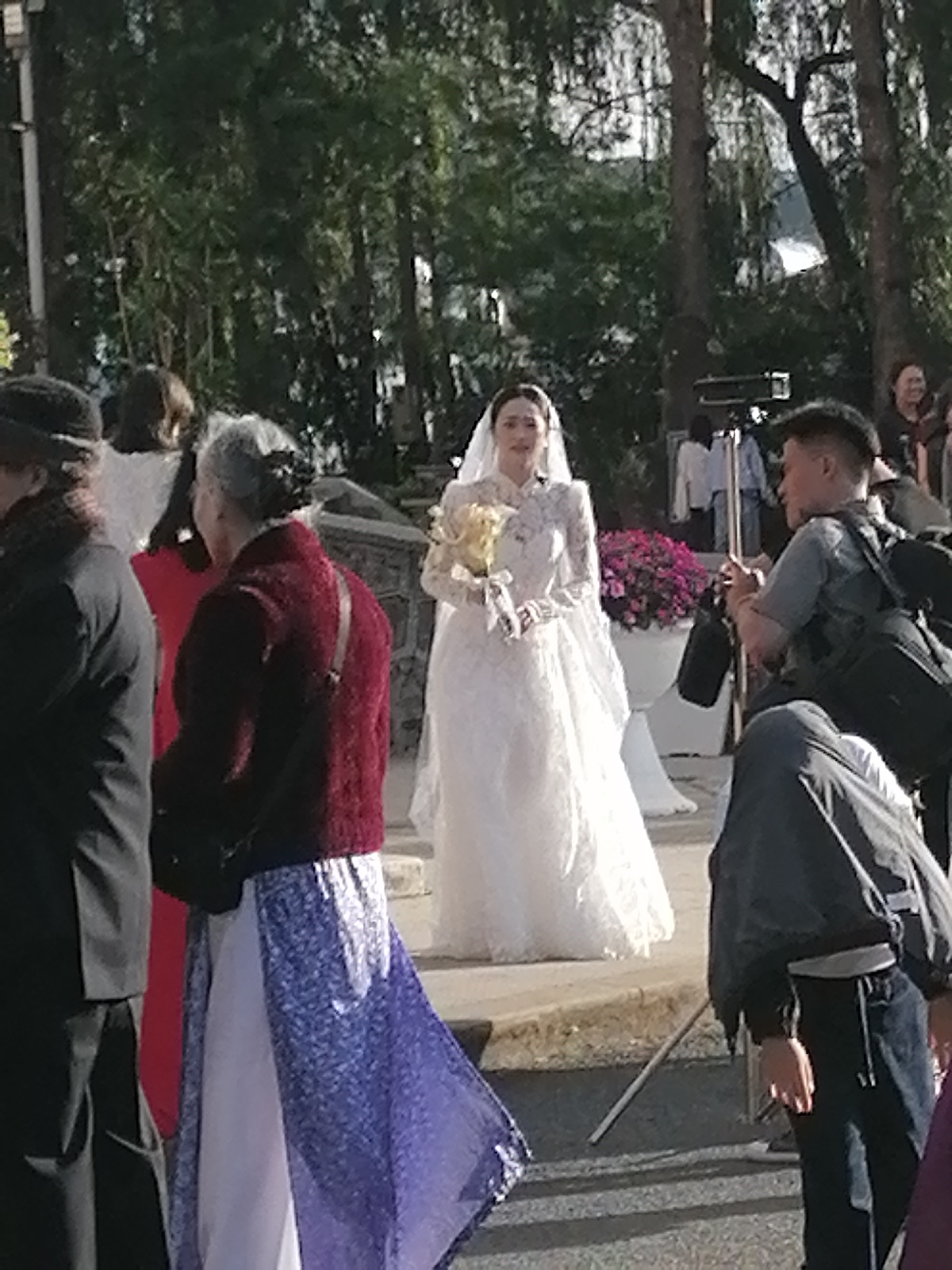 The image depicts a wedding scene in Da Lat, Vietnam, likely taking place outdoors near a natural setting given the presence of trees. In the center, a bride in a white lace wedding dress and veil holds a bouquet of yellow flowers. She appears to be looking slightly off-camera to her right. To the bride's left, an older woman in a dark red knitted vest over a patterned blue and white skirt is visible, facing away from the camera. A person in a black suit is partially visible on the far left. To the bride's right, a man with a camera bag slung over his shoulder is actively filming or photographing her. He is wearing a grey shirt and dark pants. Another person in a dark jacket and pants is also visible to the right, facing away from the camera. In the background, a white planter filled with pink flowers is situated near a white structure, possibly a wall or a pedestal. The presence of multiple people, some with professional photography equipment, suggests an organized event. The overall atmosphere appears to be one of celebration and documentation of the wedding.