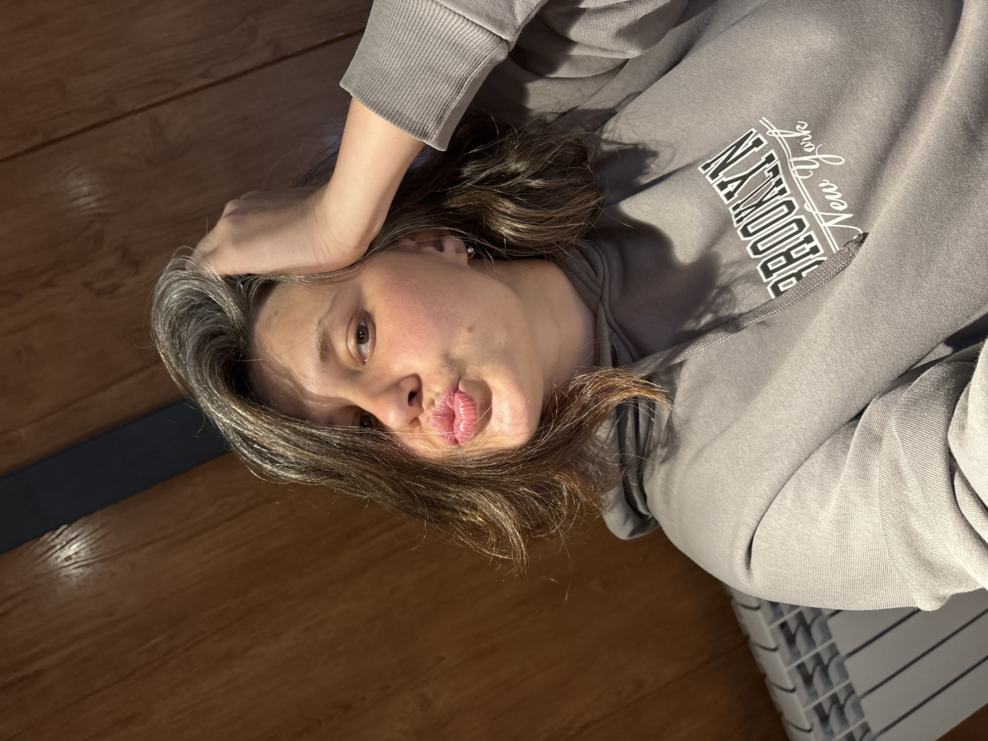A young woman with shoulder-length, dark brown hair, streaked with grey, is pictured in a close-up, selfie-style shot. She has her right hand raised and resting on the back of her head, while her lips are puckered, as if for a kiss. Her head is tilted slightly to her left. She is wearing a grey hooded sweatshirt with the words "BROOKLYN New York" printed in white lettering on the front. The background appears to be a wooden paneled wall. To the left of the frame, a white radiator is visible. The lighting suggests an indoor setting. The woman is looking directly at the camera.