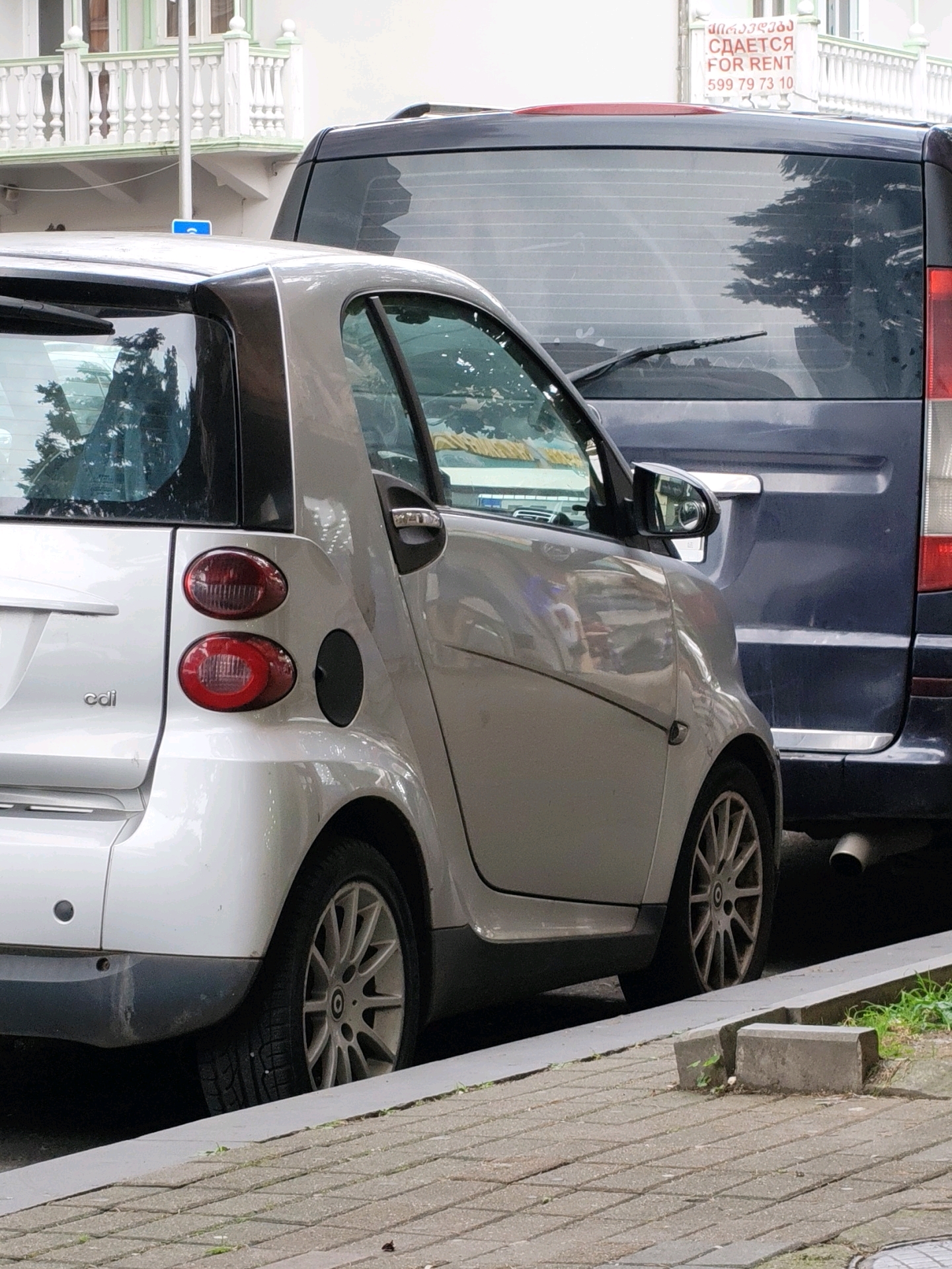 Two cars are parked on a paved street in Batumi, Georgia, with a building in the background. The car in the foreground is a silver Smart Fortwo, parked parallel to the curb. The car behind it is a dark blue van, possibly a Mercedes-Benz Vito or similar model. A sign advertising "FOR RENT" in Russian and Georgian is visible on the building behind the van. The street is lined with a sidewalk and a low concrete curb. The scene appears to be a typical urban streetscape.